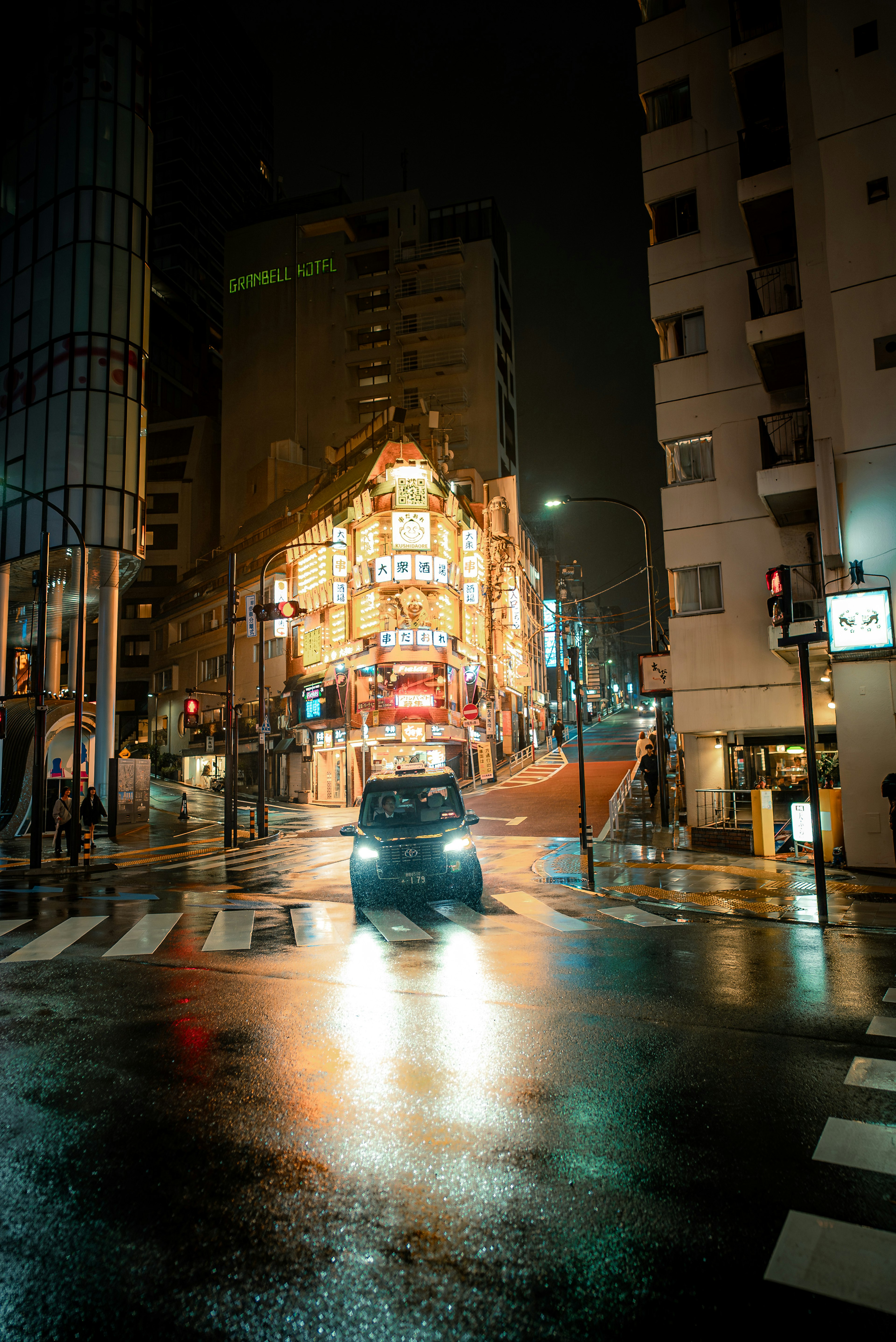 Un coche en una intersección urbana por la noche con luces de neón reflejándose en el pavimento mojado