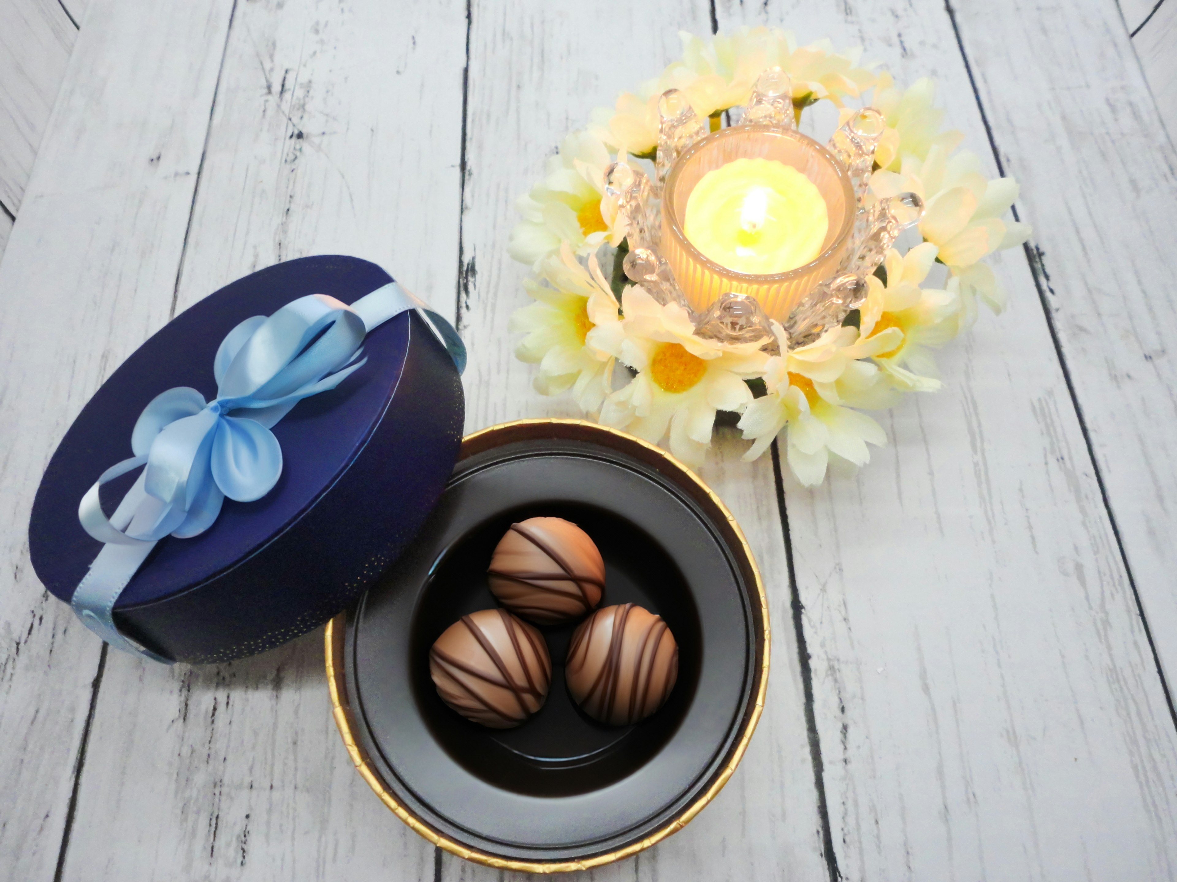 Une boîte cadeau avec un ruban bleu contenant des boules en chocolat sur une assiette décorative entourée d'une bougie et d'un arrangement floral