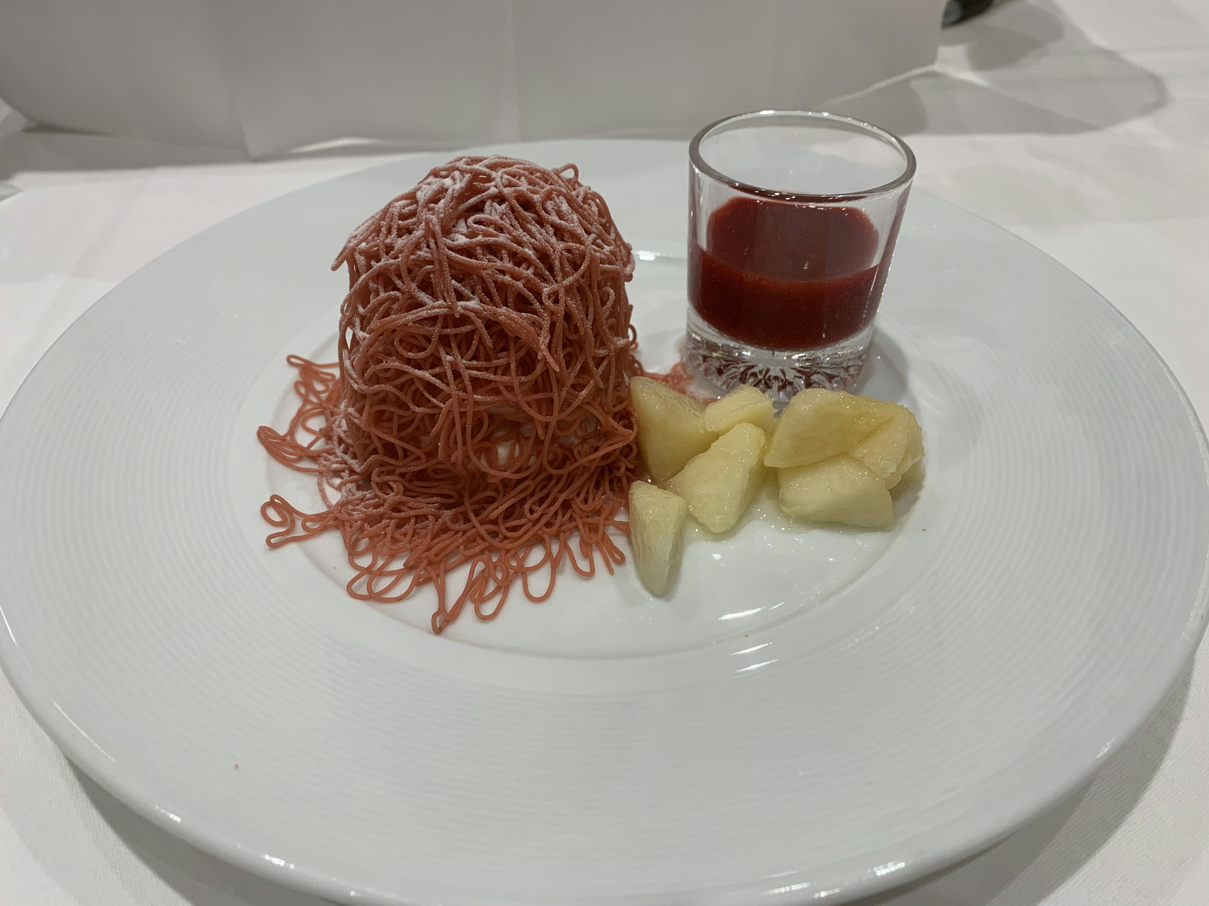 Dessert su un piatto bianco con crema rosa a forma di noodle un piccolo bicchiere di salsa rossa e fette di mela