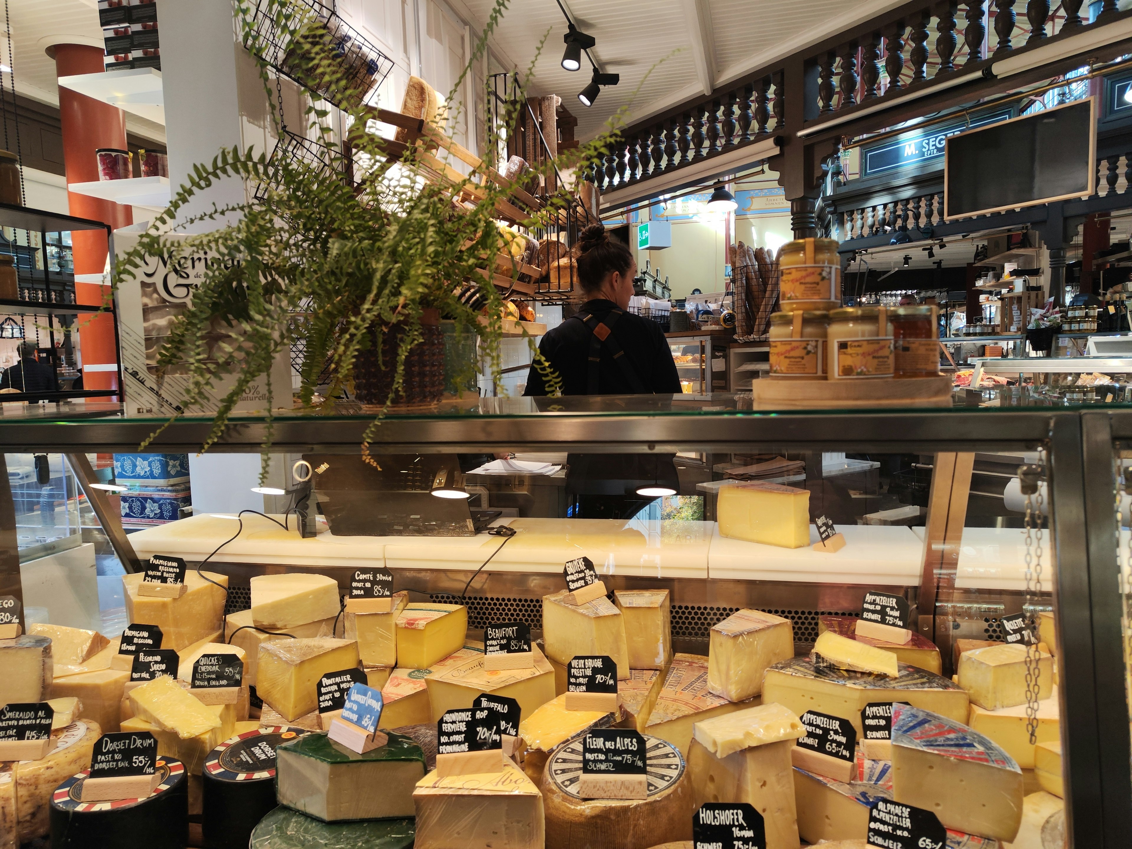 Variety of cheeses displayed in a showcase with a shop attendant in the background