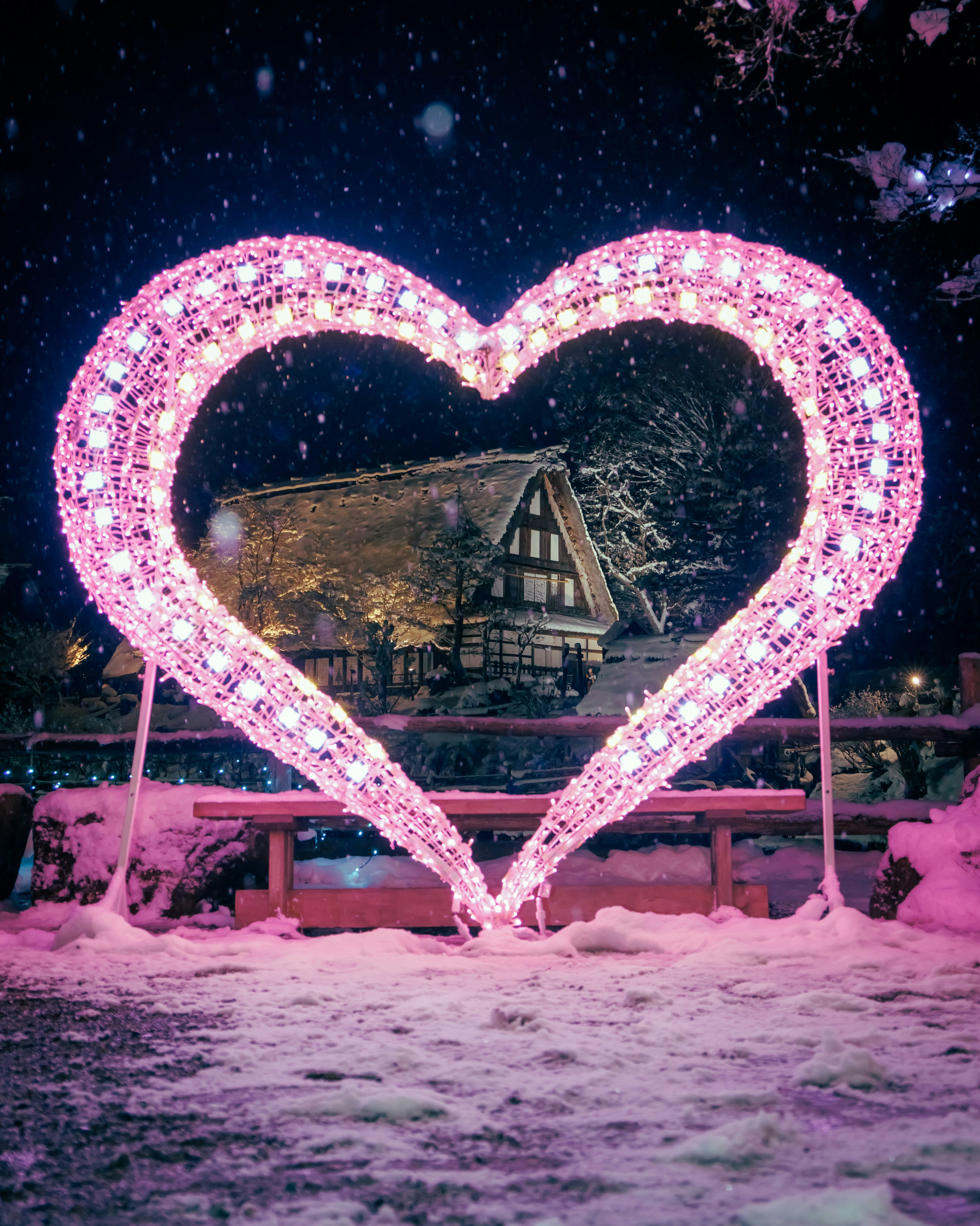 Installazione luminosa a forma di cuore rosa nella neve con una casa sullo sfondo