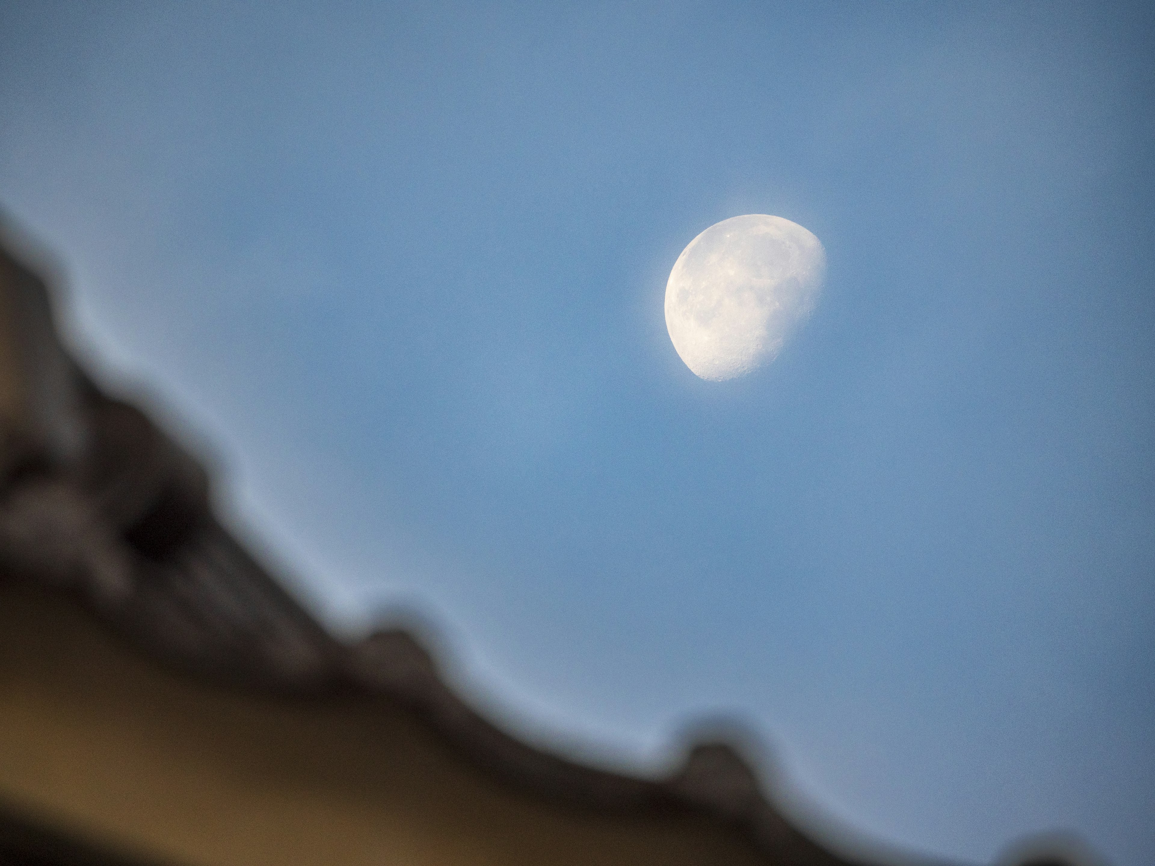 Photo of a half moon in the blue sky with part of a roof visible