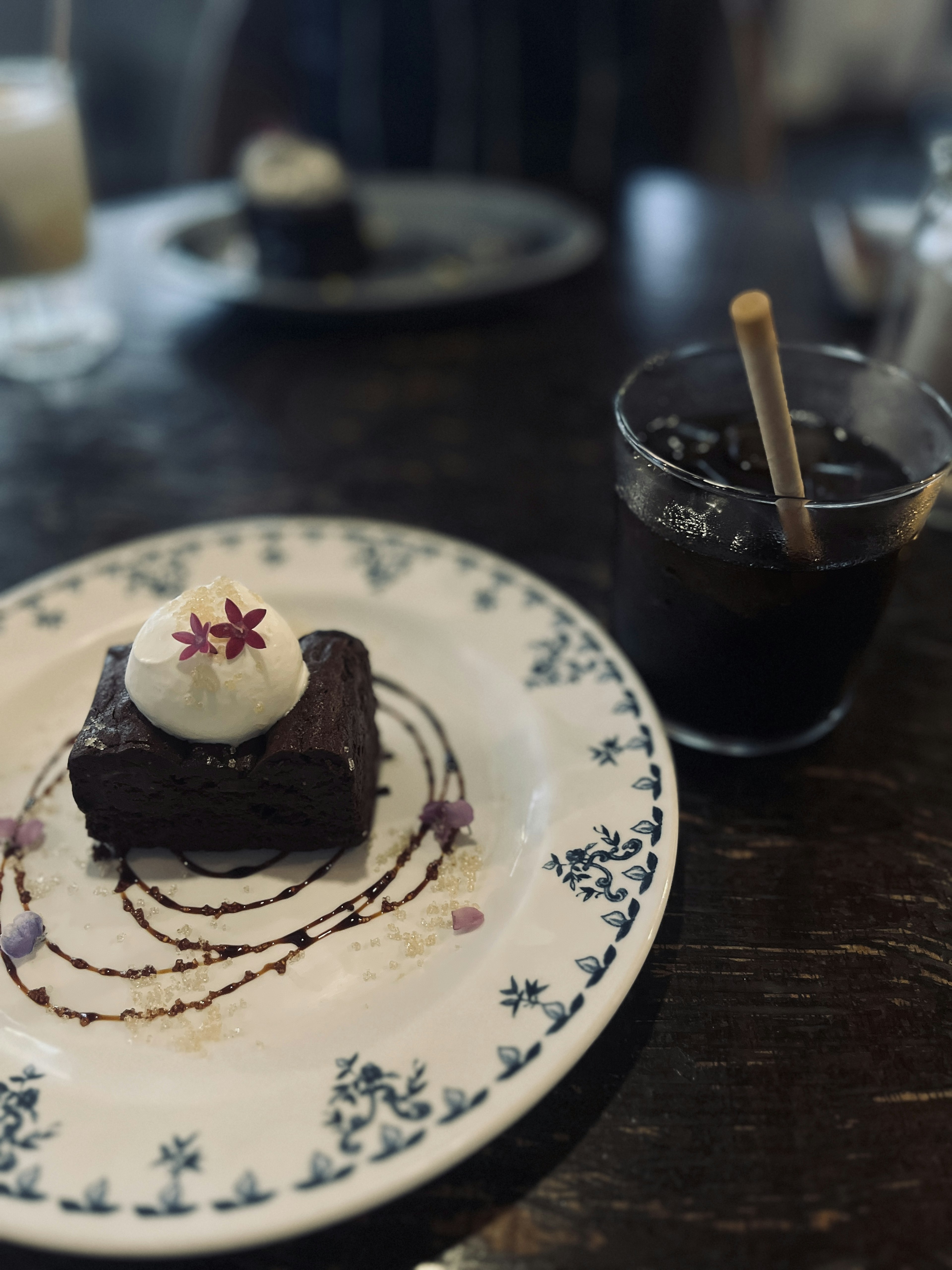 Chocolate cake on a decorative plate with cream and a flower garnish alongside a glass of iced drink