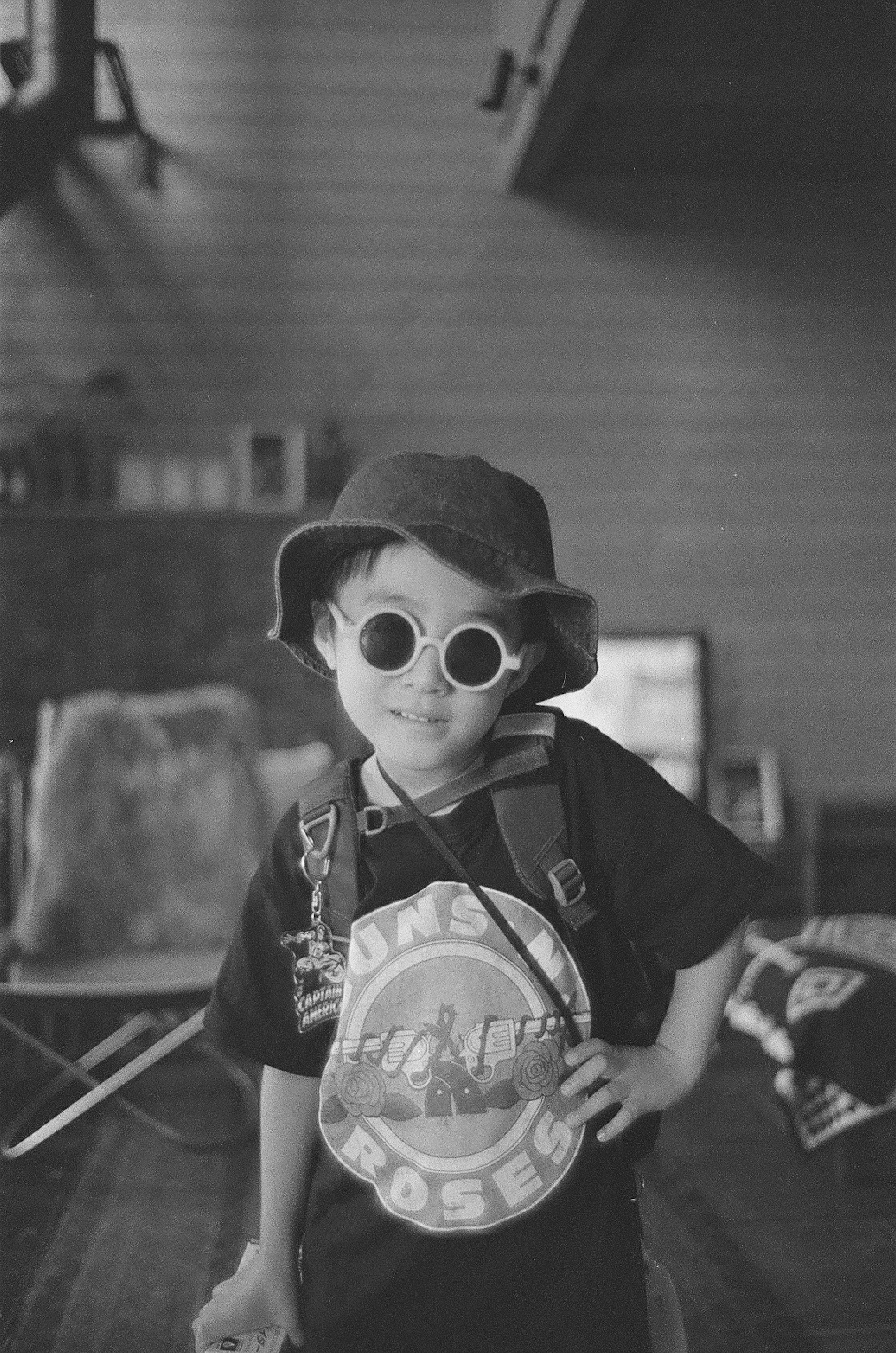 Black and white photo of a child wearing a hat and sunglasses holding a backpack