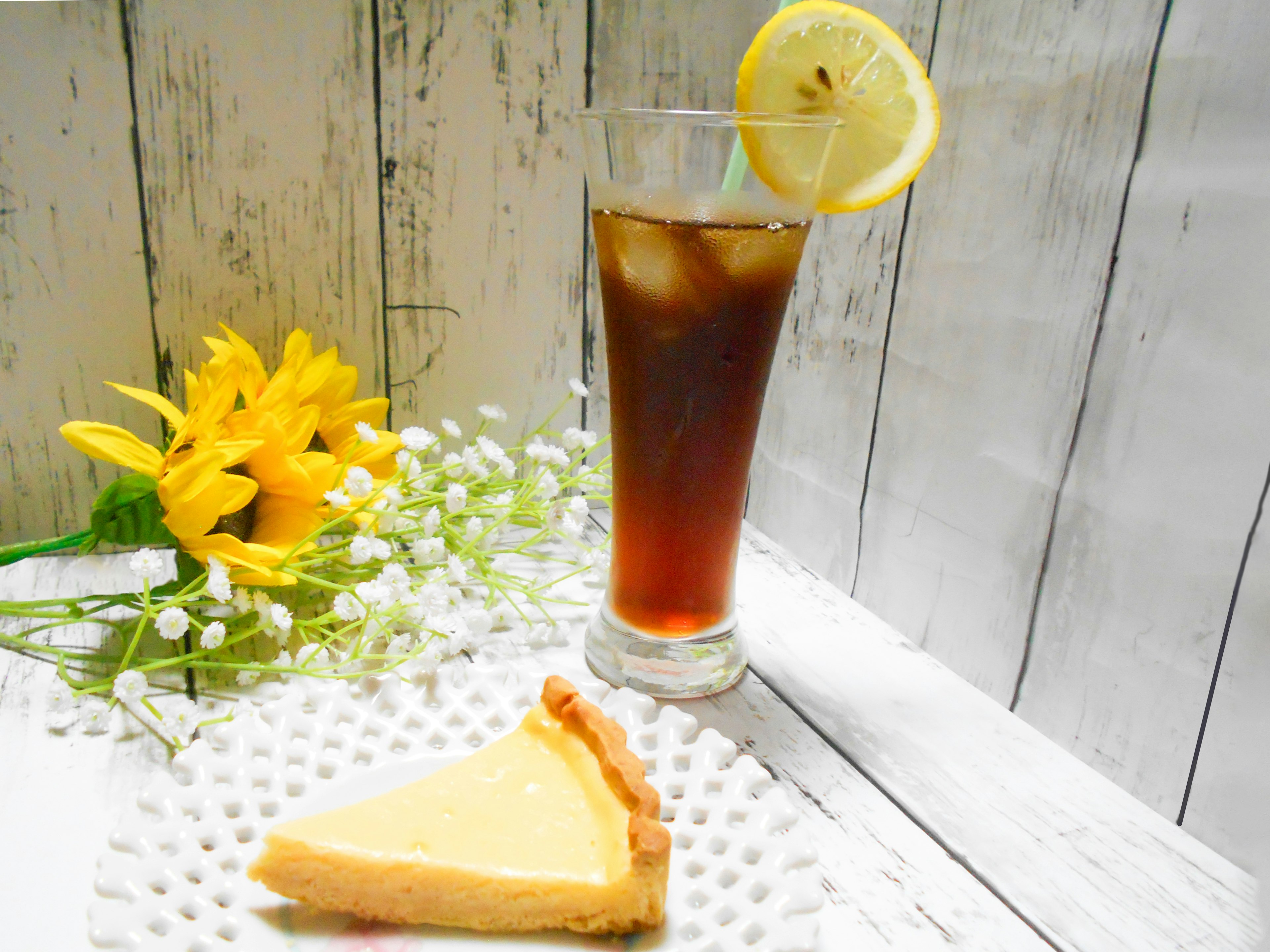 Segelas es teh dengan lemon dan sepotong cheesecake di samping susunan bunga kuning