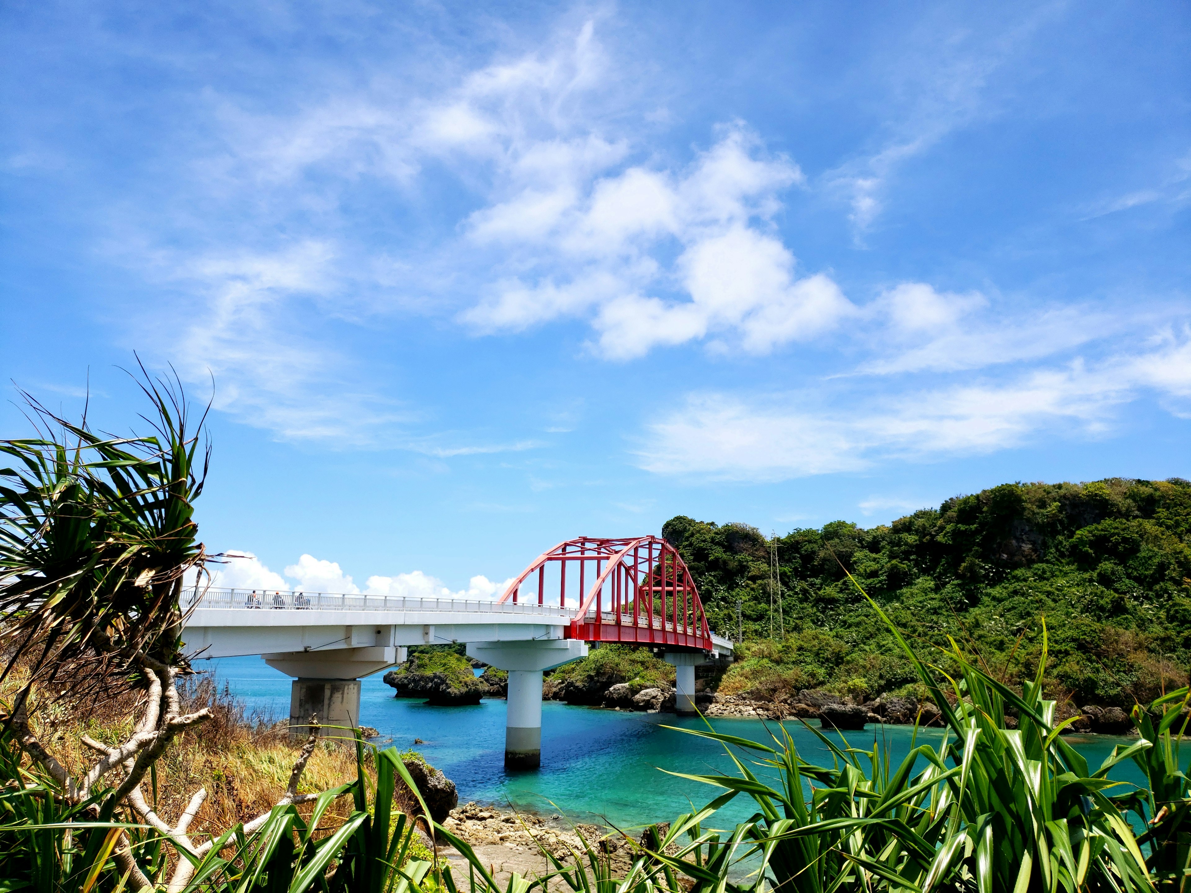 A scenic view featuring a red bridge over clear blue water surrounded by green hills and a bright sky