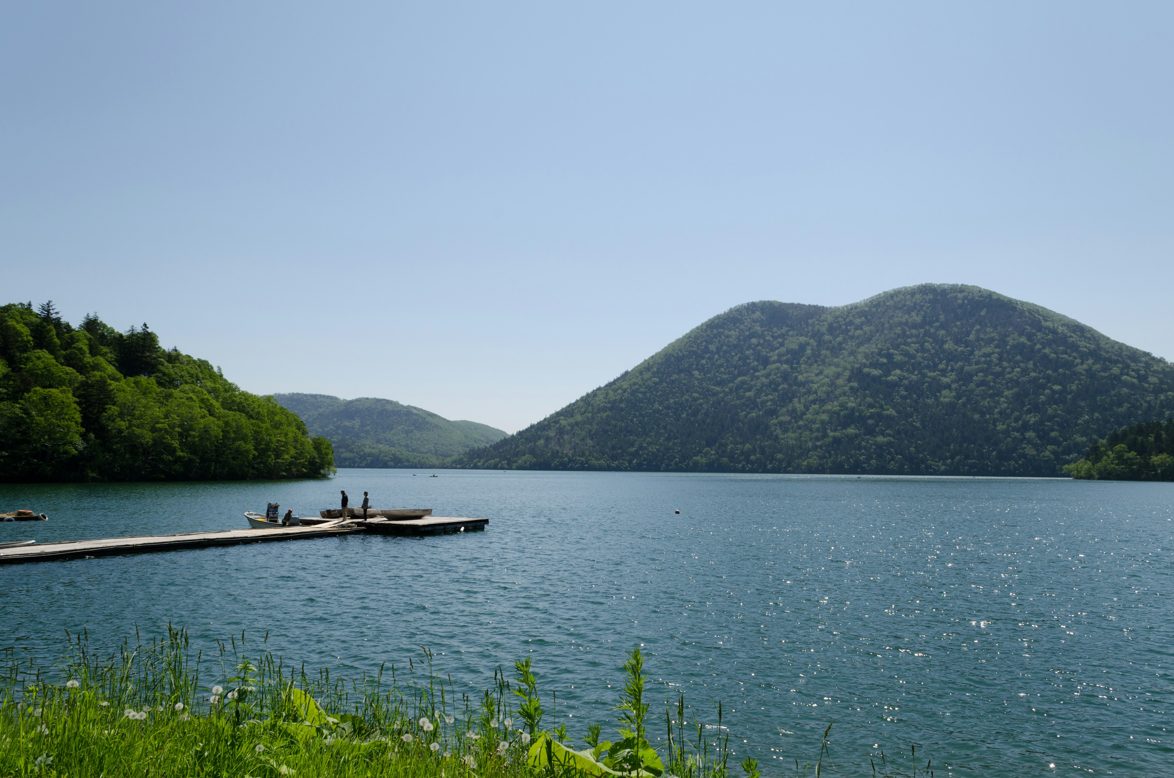 Scenic view of a calm lake and mountains under a clear blue sky with green trees and people on a dock