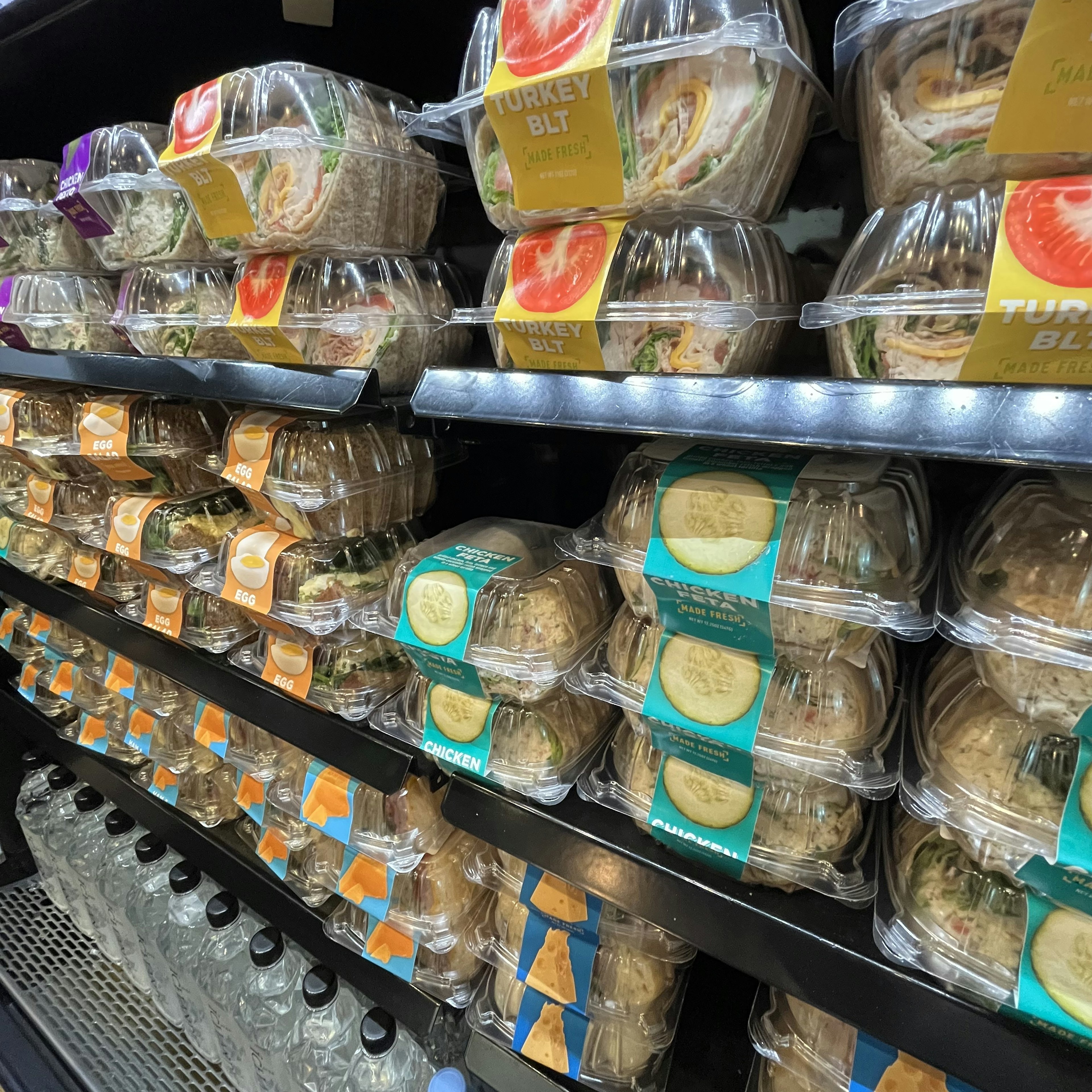 Refrigerator shelf displaying packaged sandwiches and bottled drinks