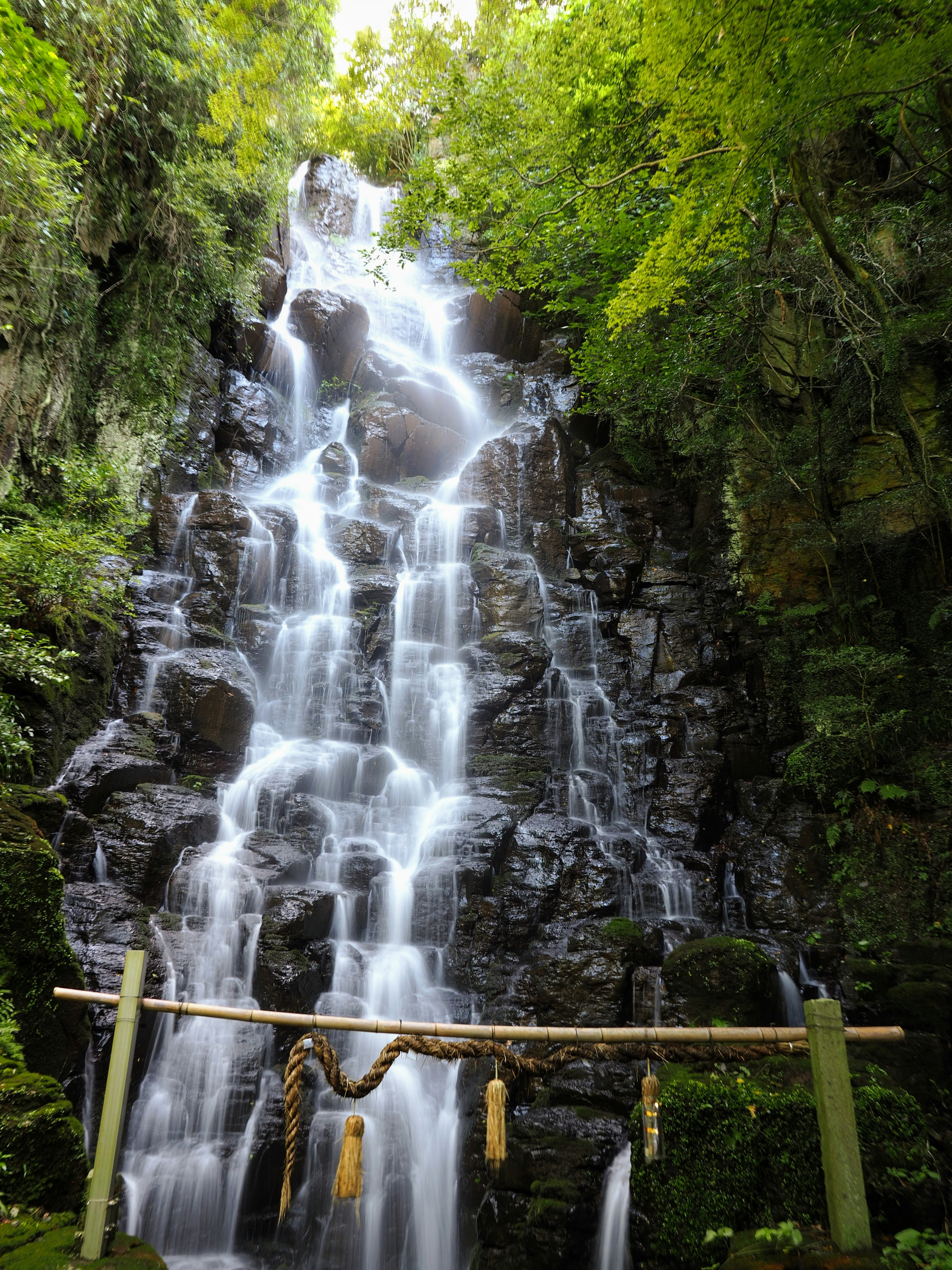 Gambar air terjun yang indah dikelilingi oleh vegetasi hijau subur dengan struktur kayu suci dan tali di depan