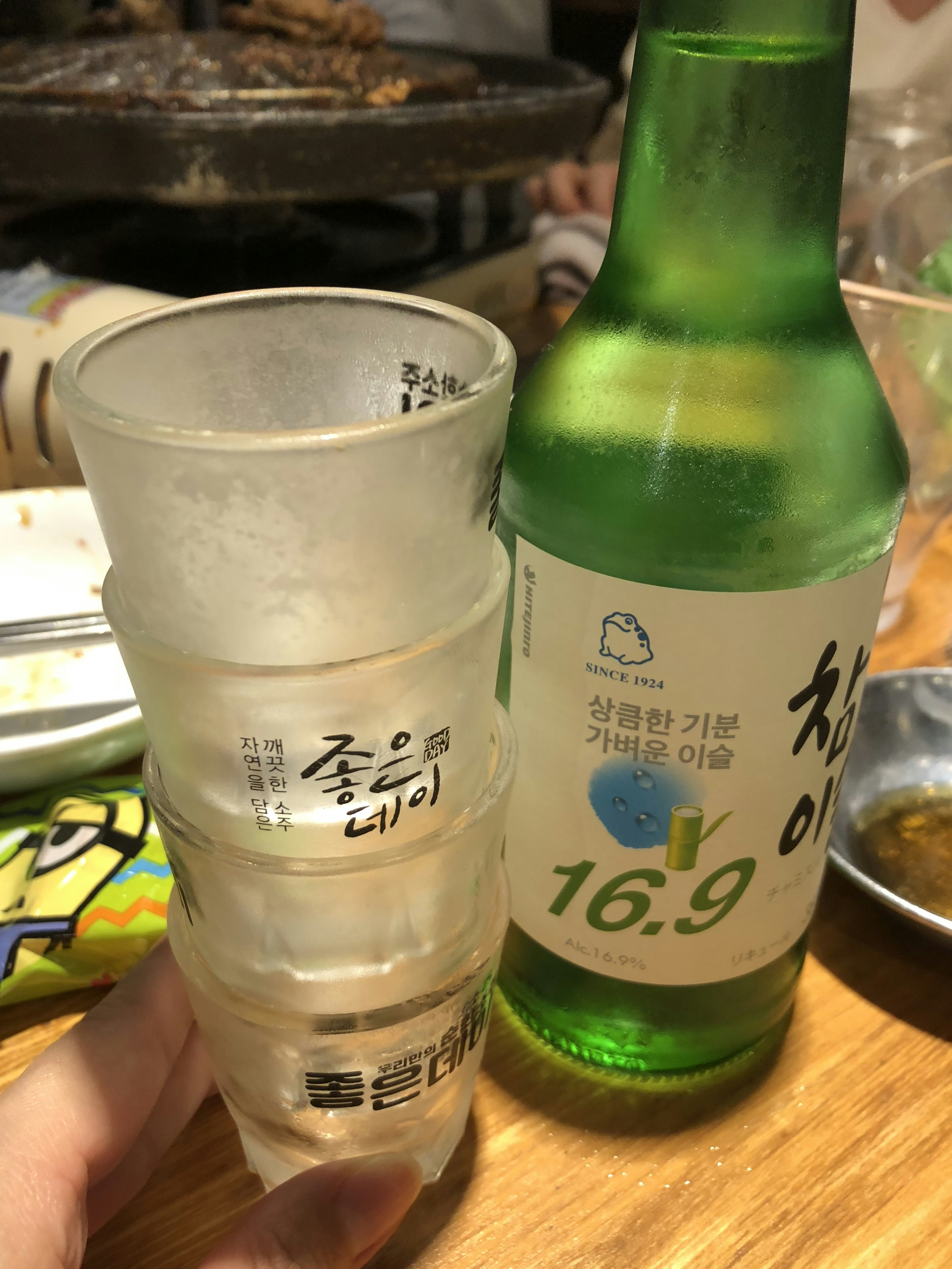 A hand holding stacked clear glasses beside a green soju bottle