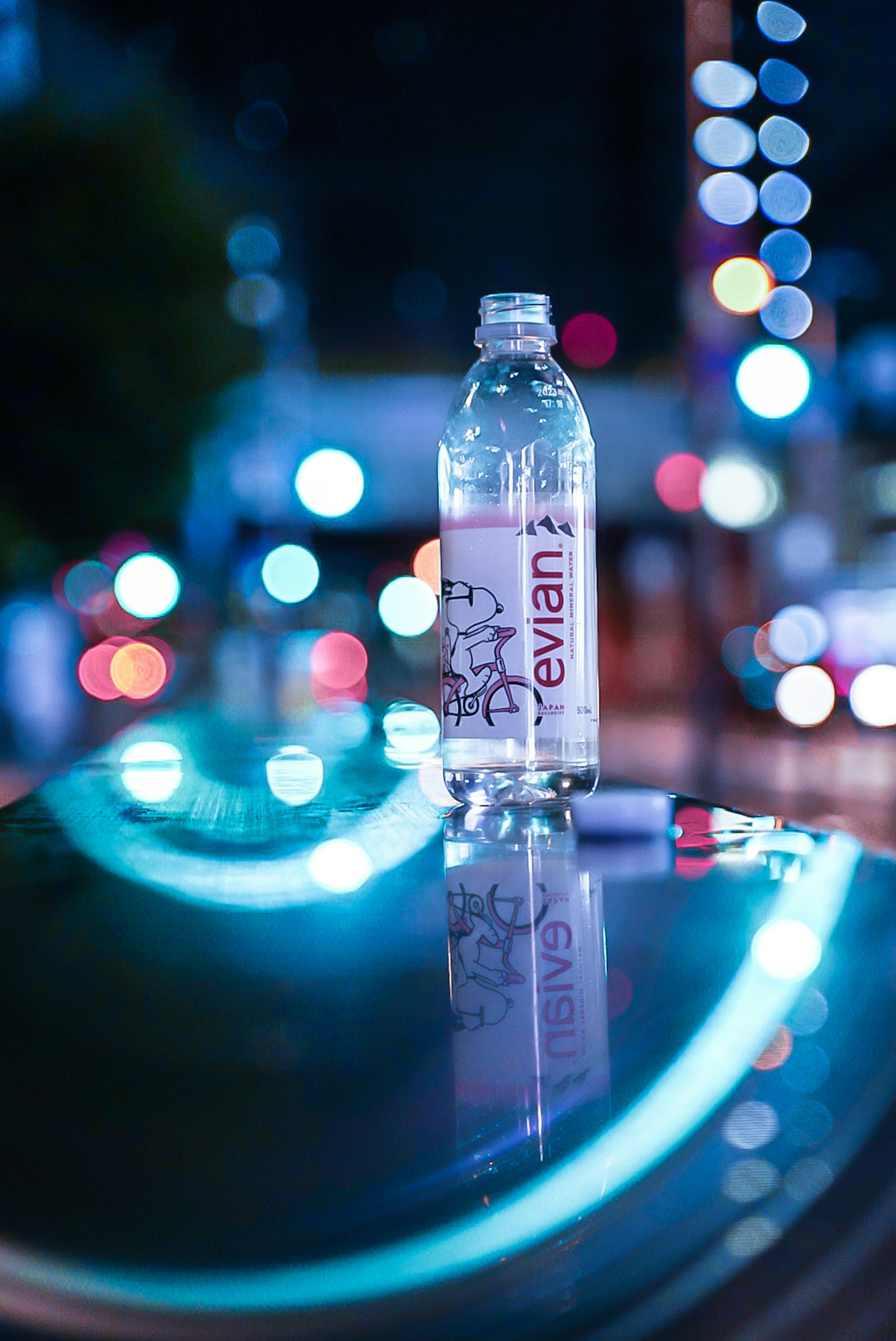 Una botella de agua transparente colocada sobre una superficie reflectante con luces de bokeh coloridas de fondo