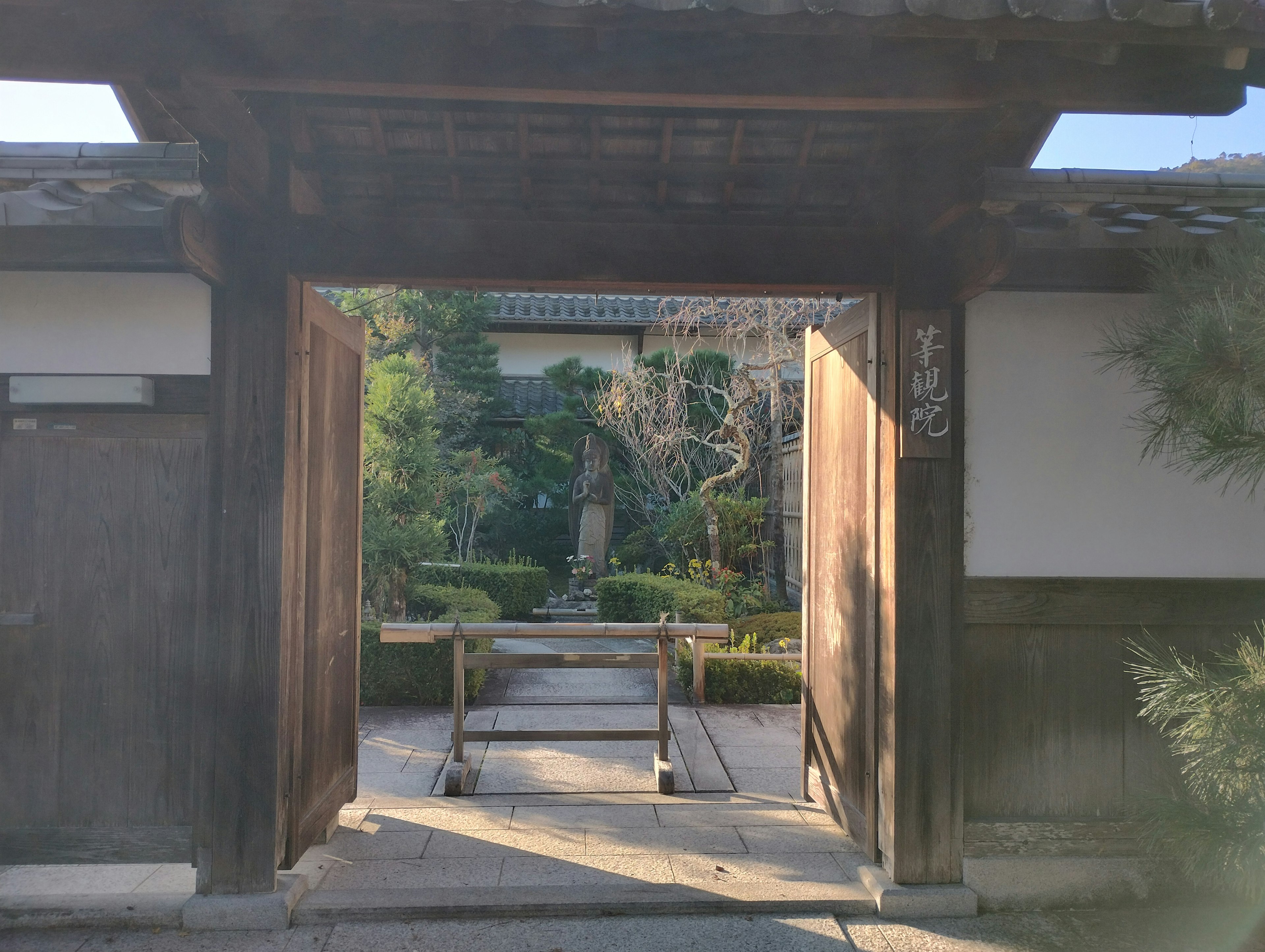 Blick auf einen japanischen Garten durch ein Holztor mit Grünpflanzen und Steinskulpturen