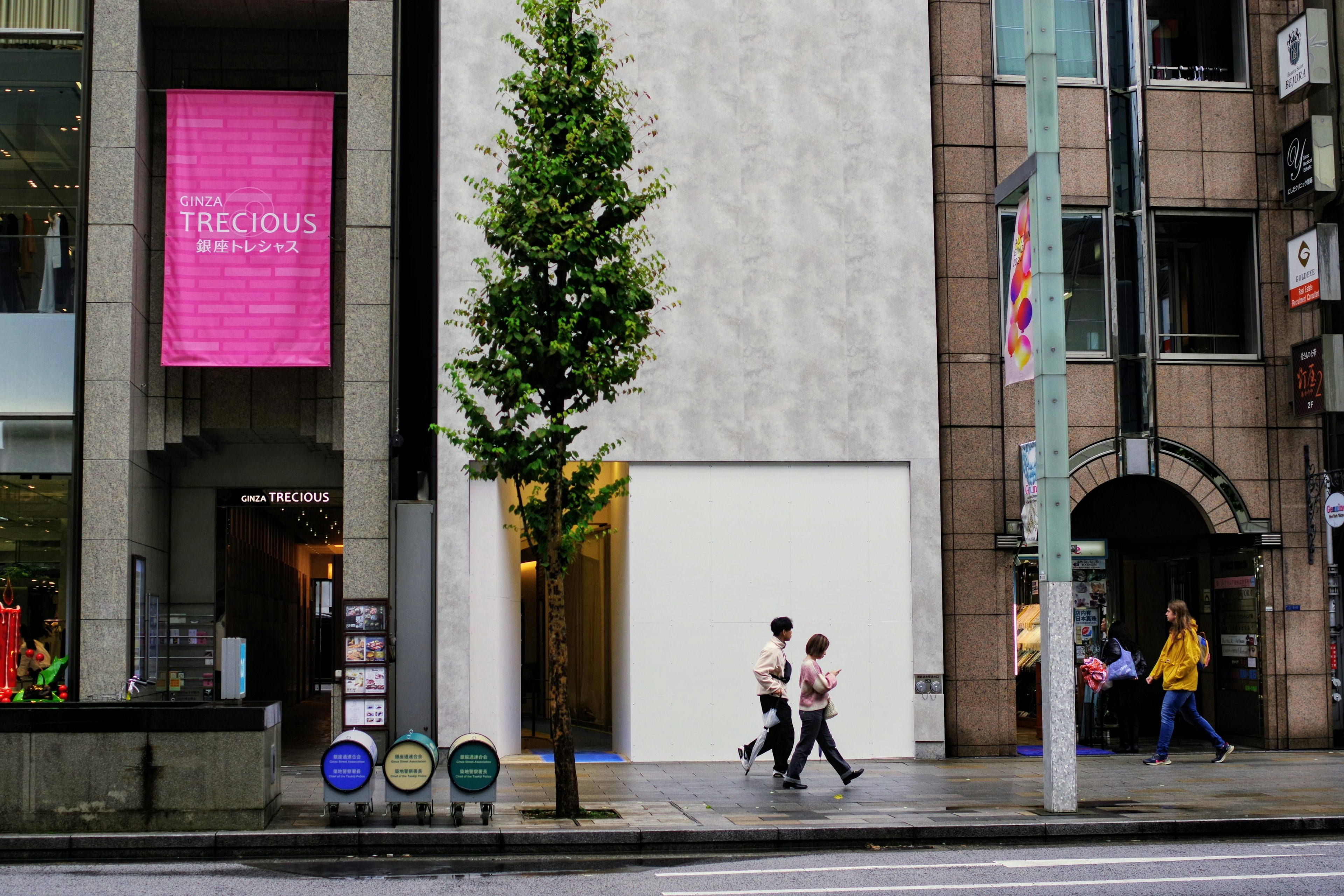 Escena urbana con personas caminando por la calle que presenta una pared blanca simple y un árbol verde