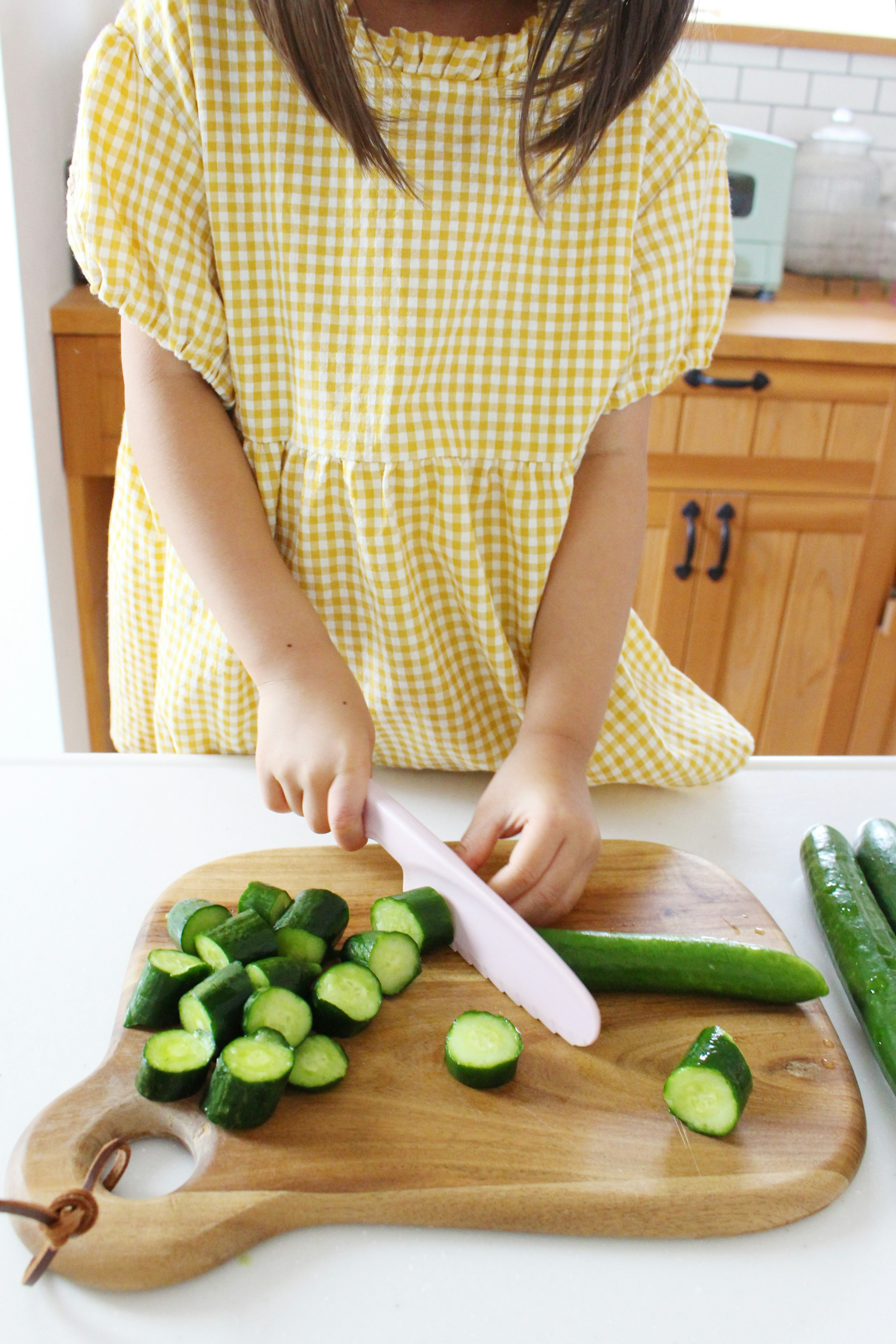 A girl in a yellow checkered dress slicing cucumbers in a kitchen