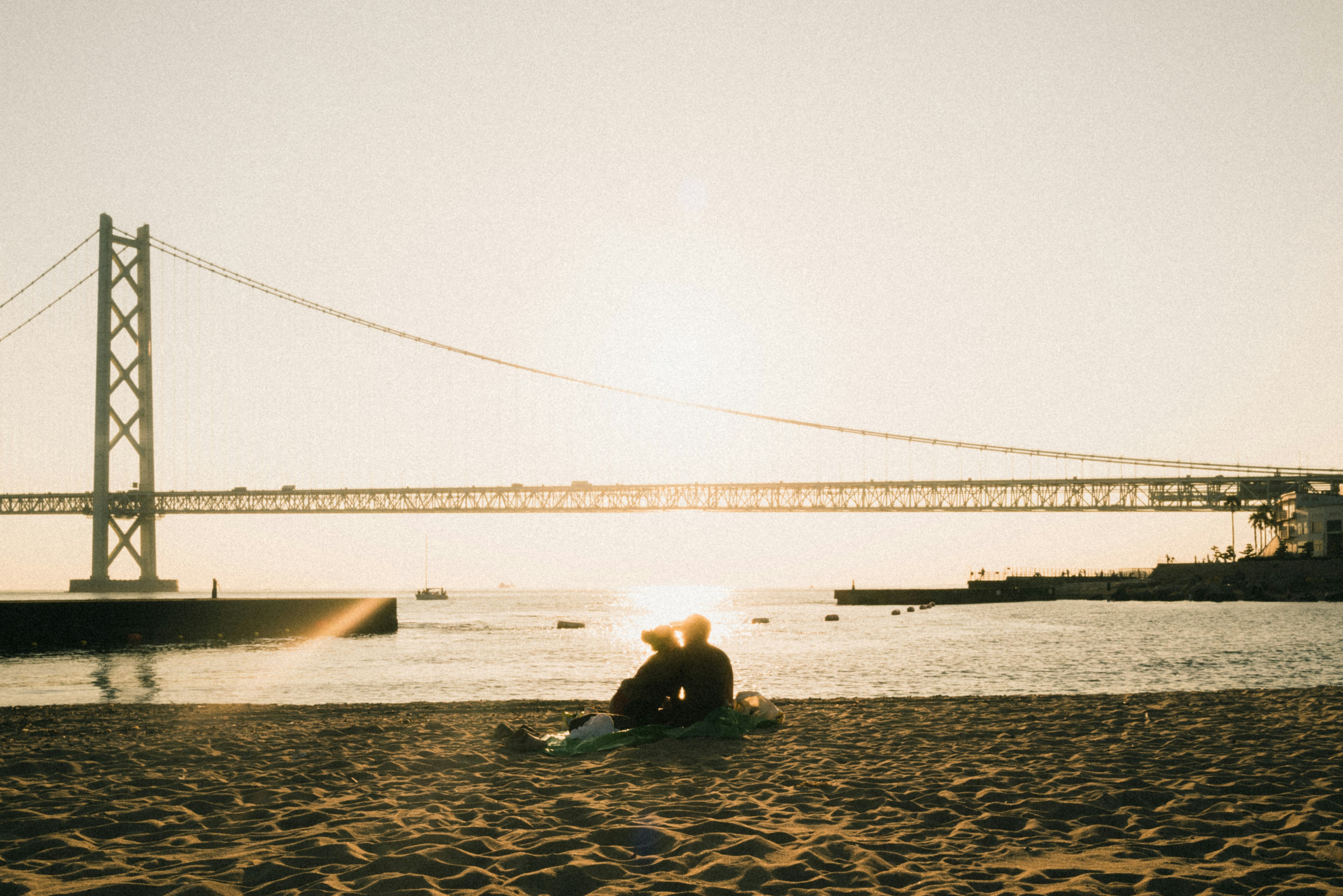 Couple sitting on the beach at sunset with a bridge in the background