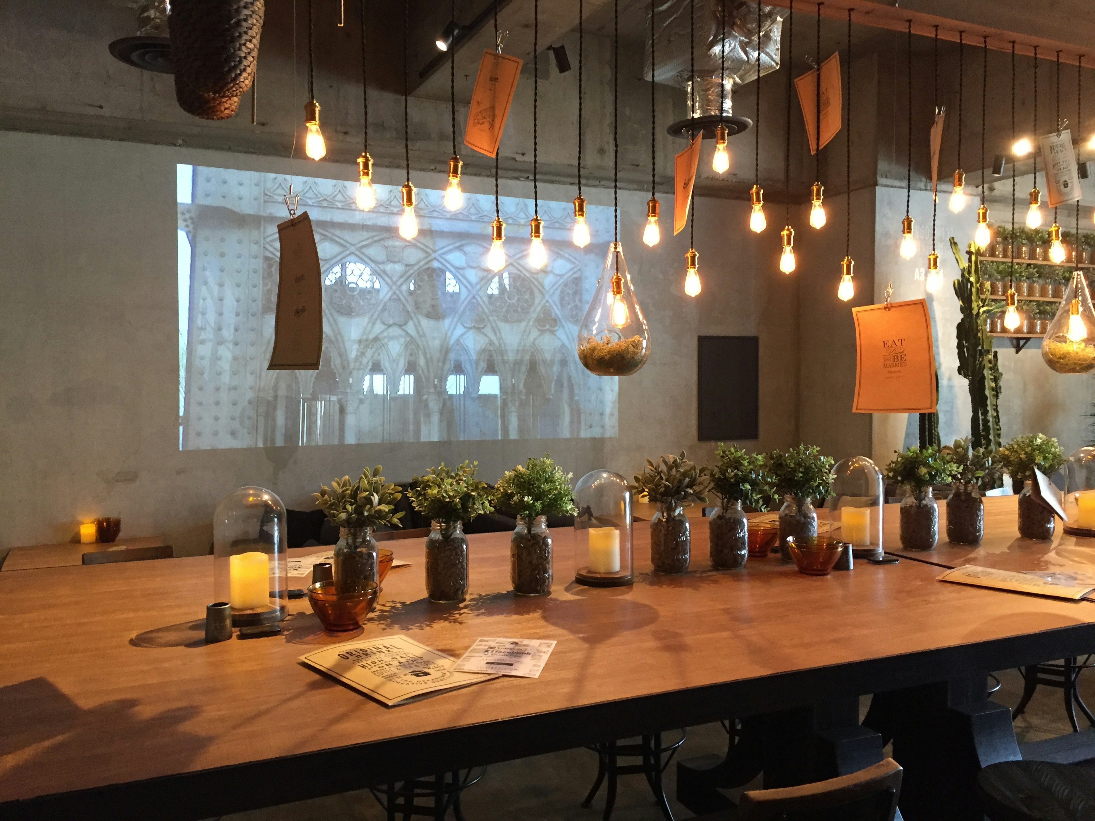 Interior de una cafetería moderna con una larga mesa de madera, luces colgantes y una imagen proyectada en la pared