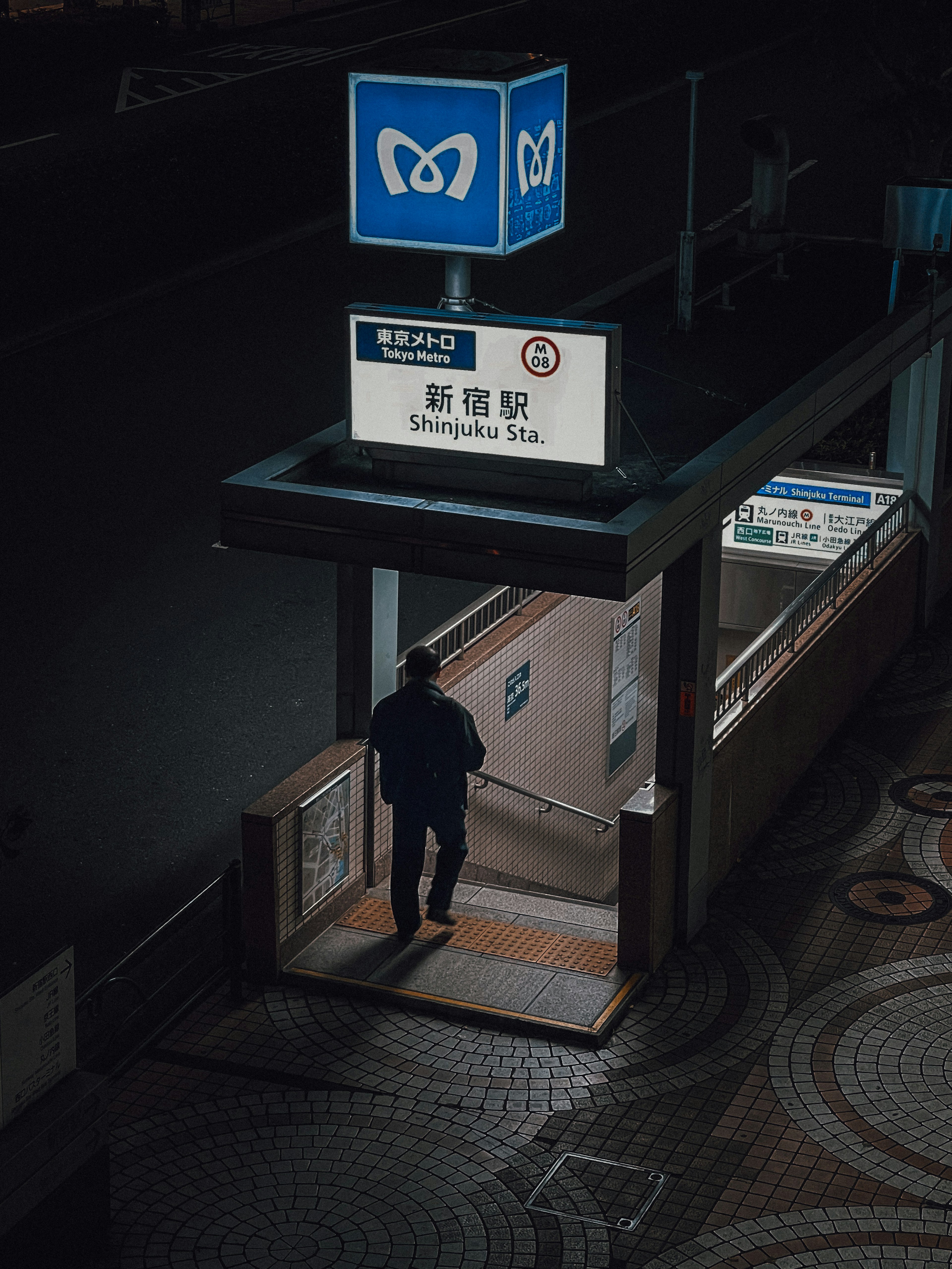 A man standing at the entrance of a subway station at night with a blue sign