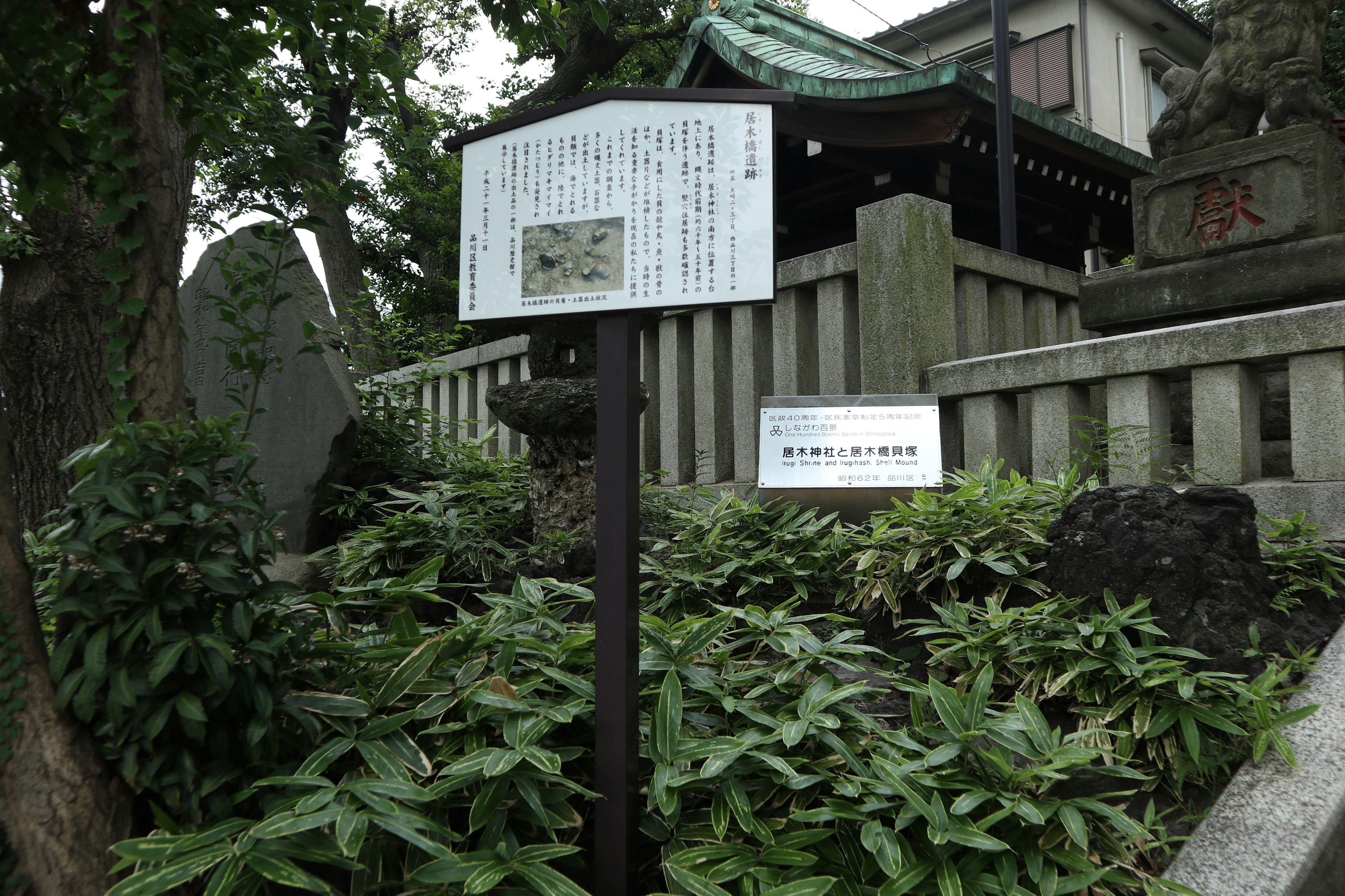 A view featuring a sign surrounded by green plants and part of a shrine building