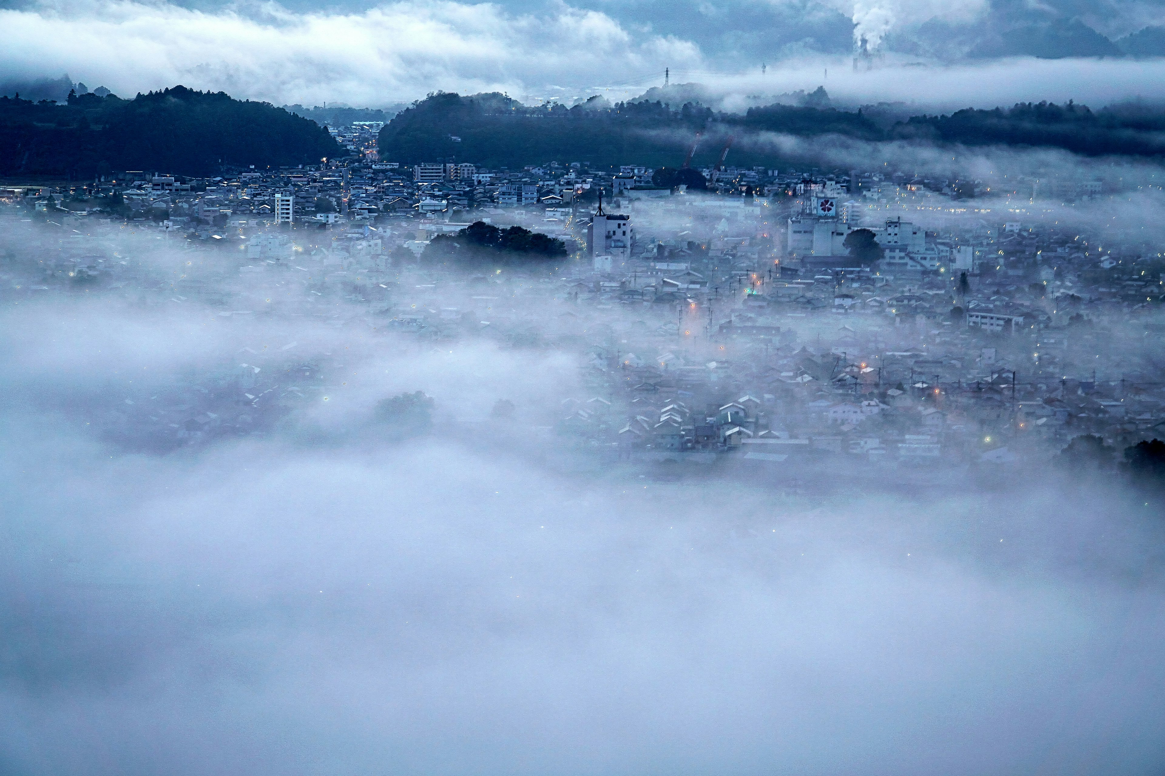 Cityscape shrouded in fog with distant mountains and city lights peeking through clouds