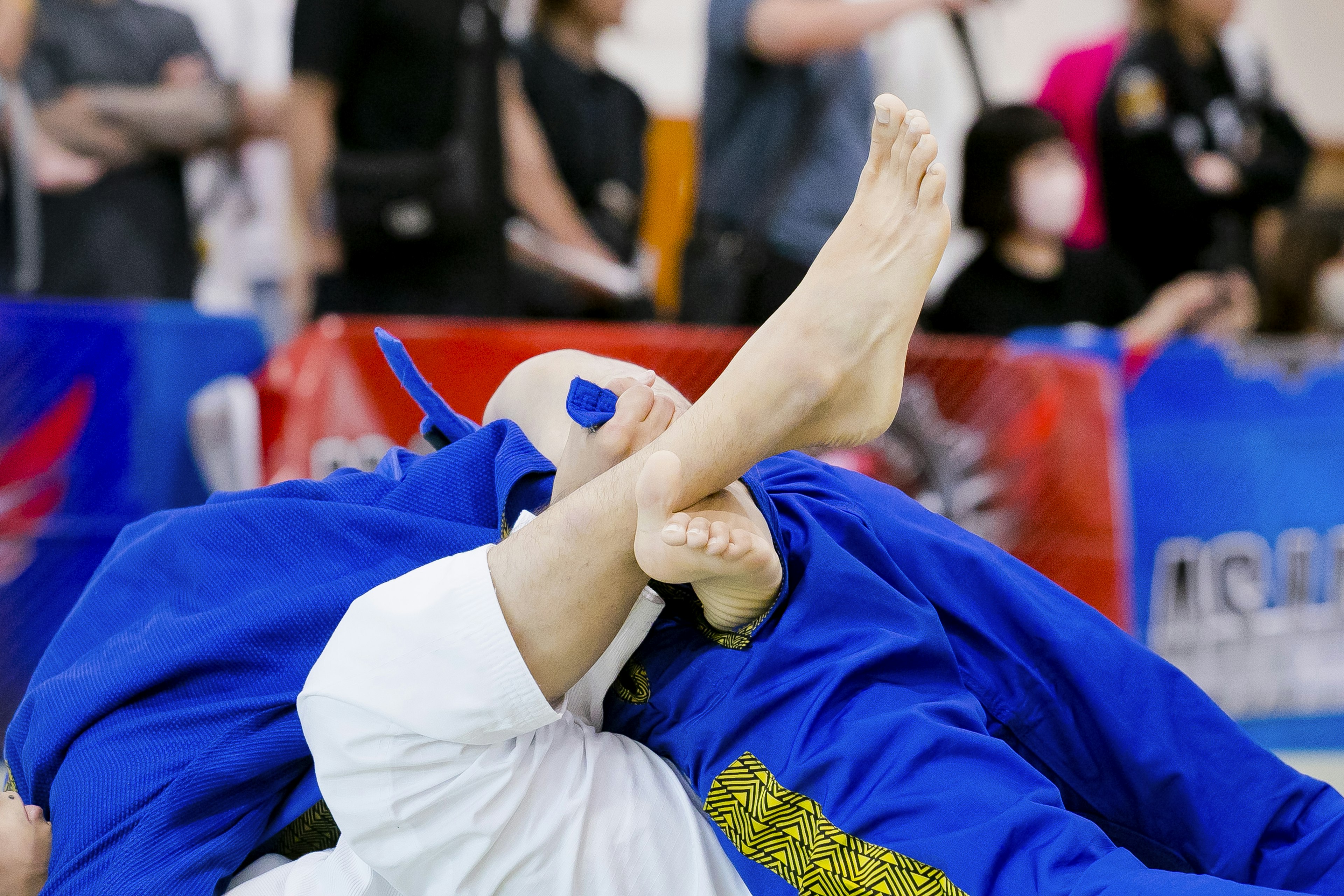 Two judo competitors are engaged in a match with the blue gi competitor entangling the legs of the white gi competitor