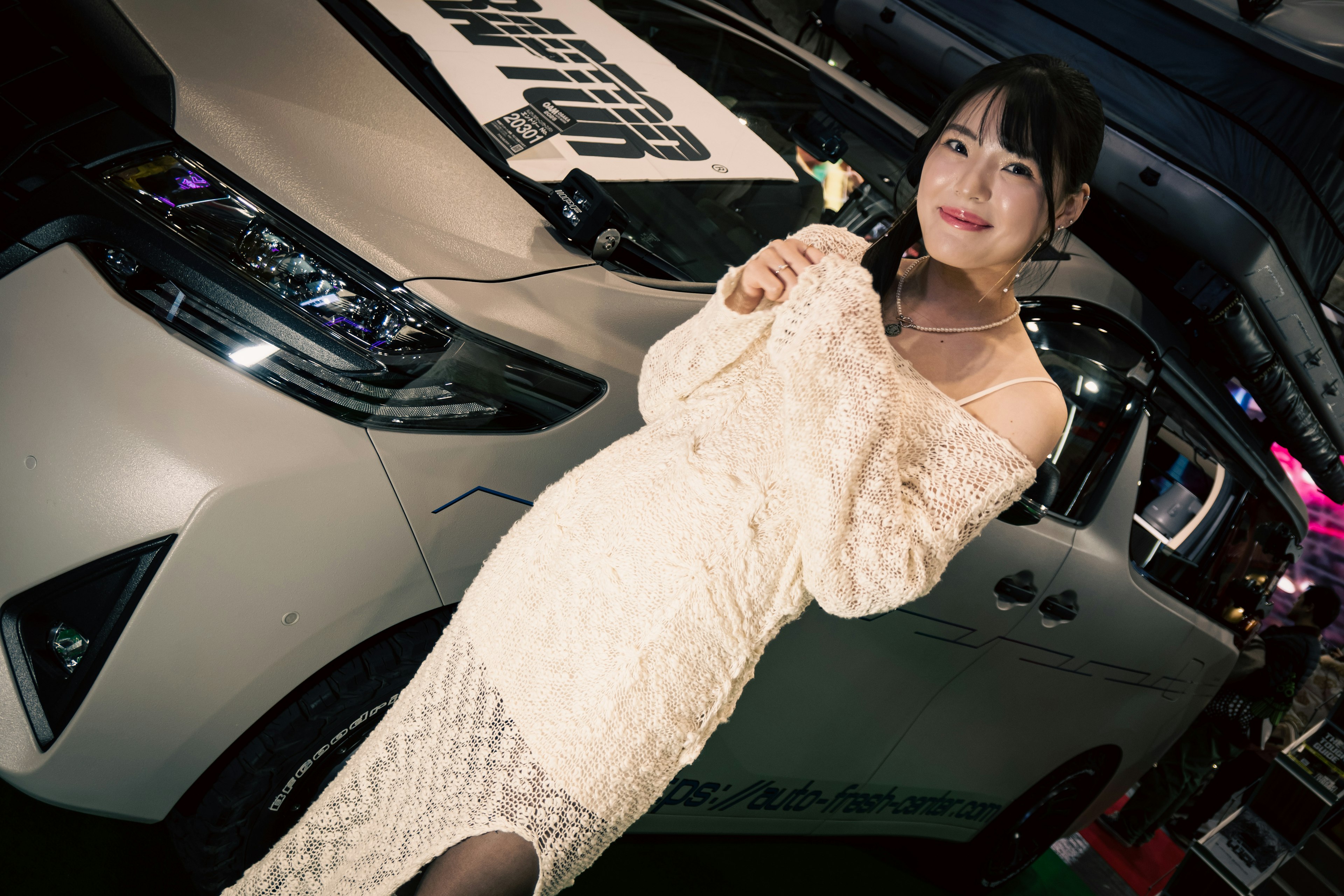 A woman in a white dress stands in front of a car