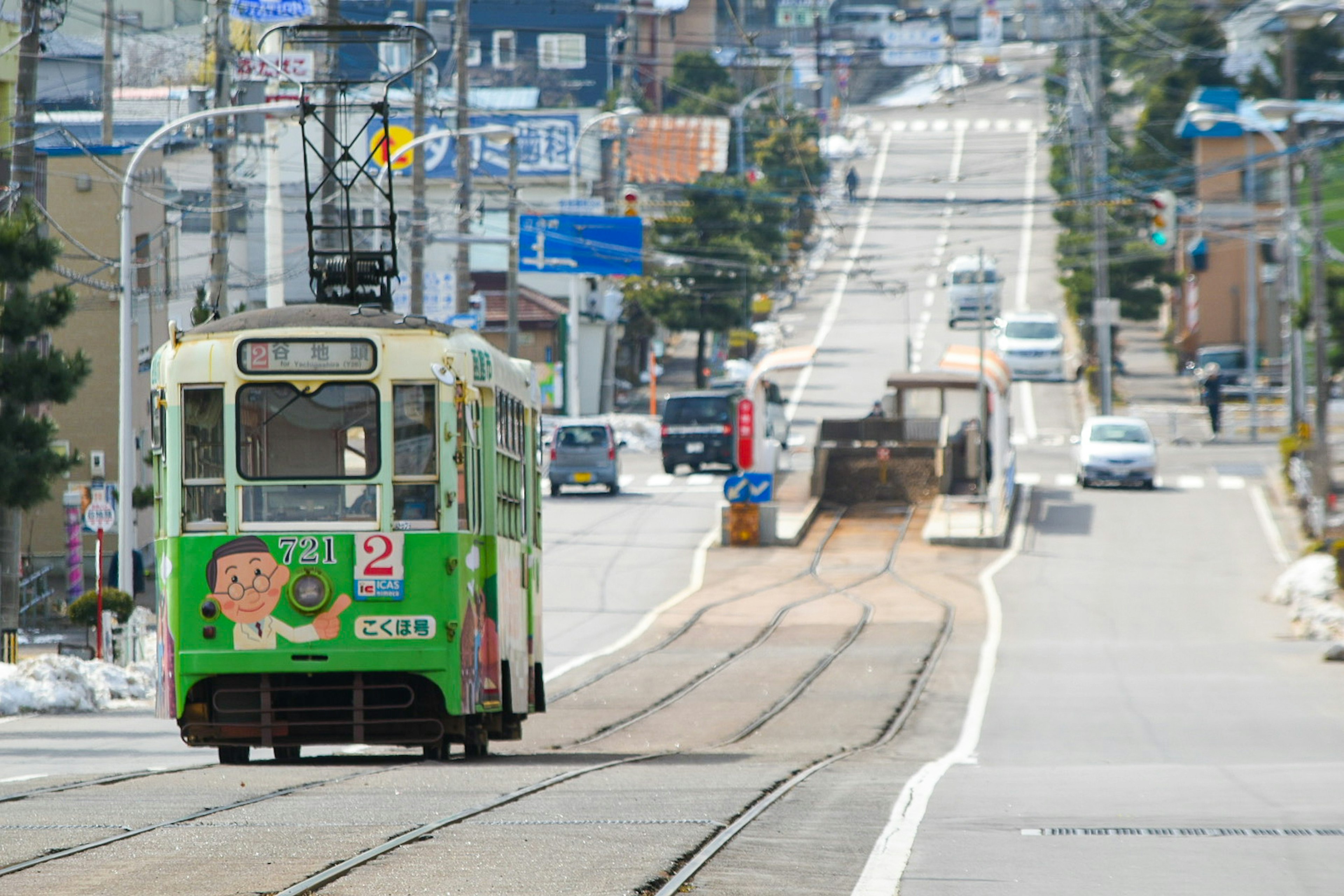 Grüne Straßenbahn fährt entlang einer Straße mit Geschäften und Fahrzeugen