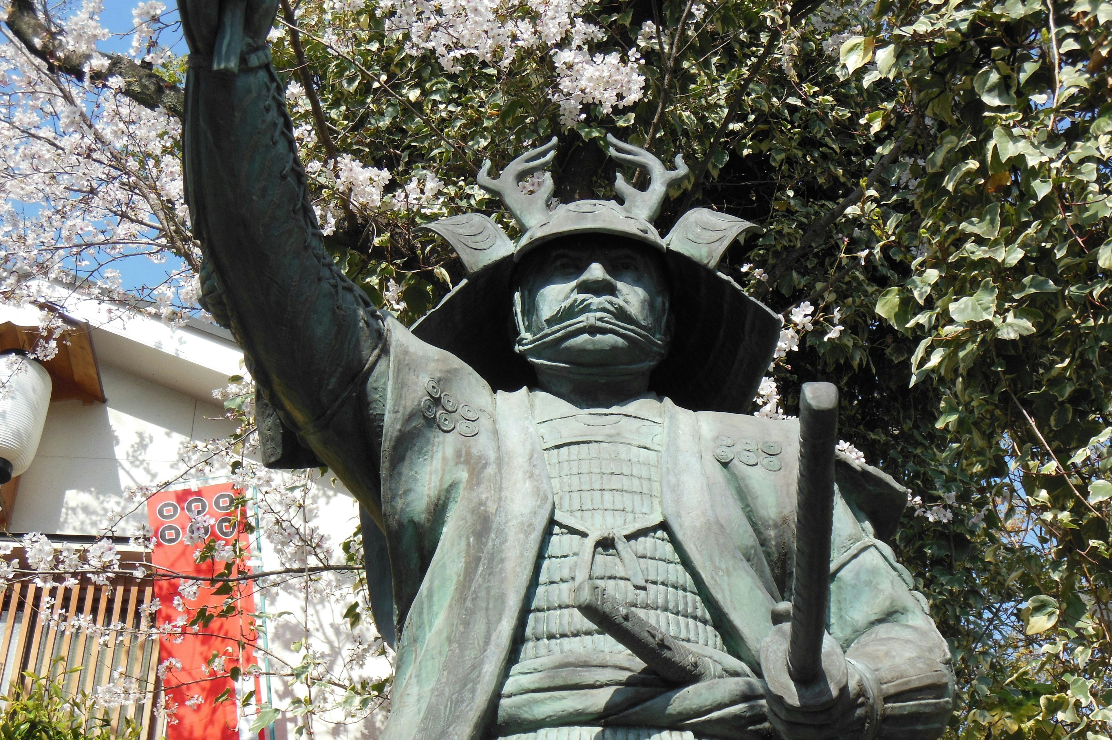 Bronze statue of a samurai standing under cherry blossom trees holding a sword raised high