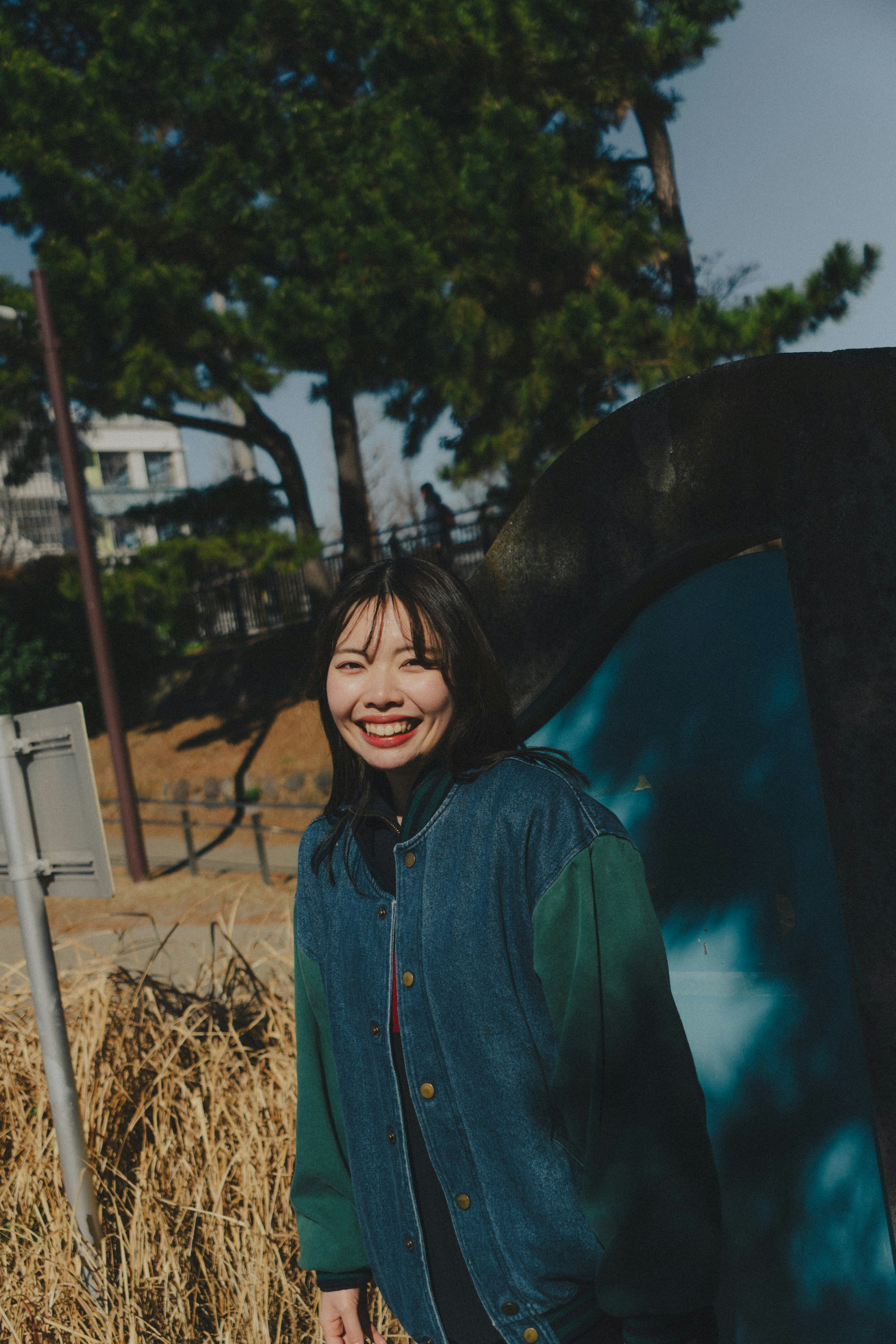 Mujer sonriente con chaqueta azul de pie frente a árboles verdes y una pared azul