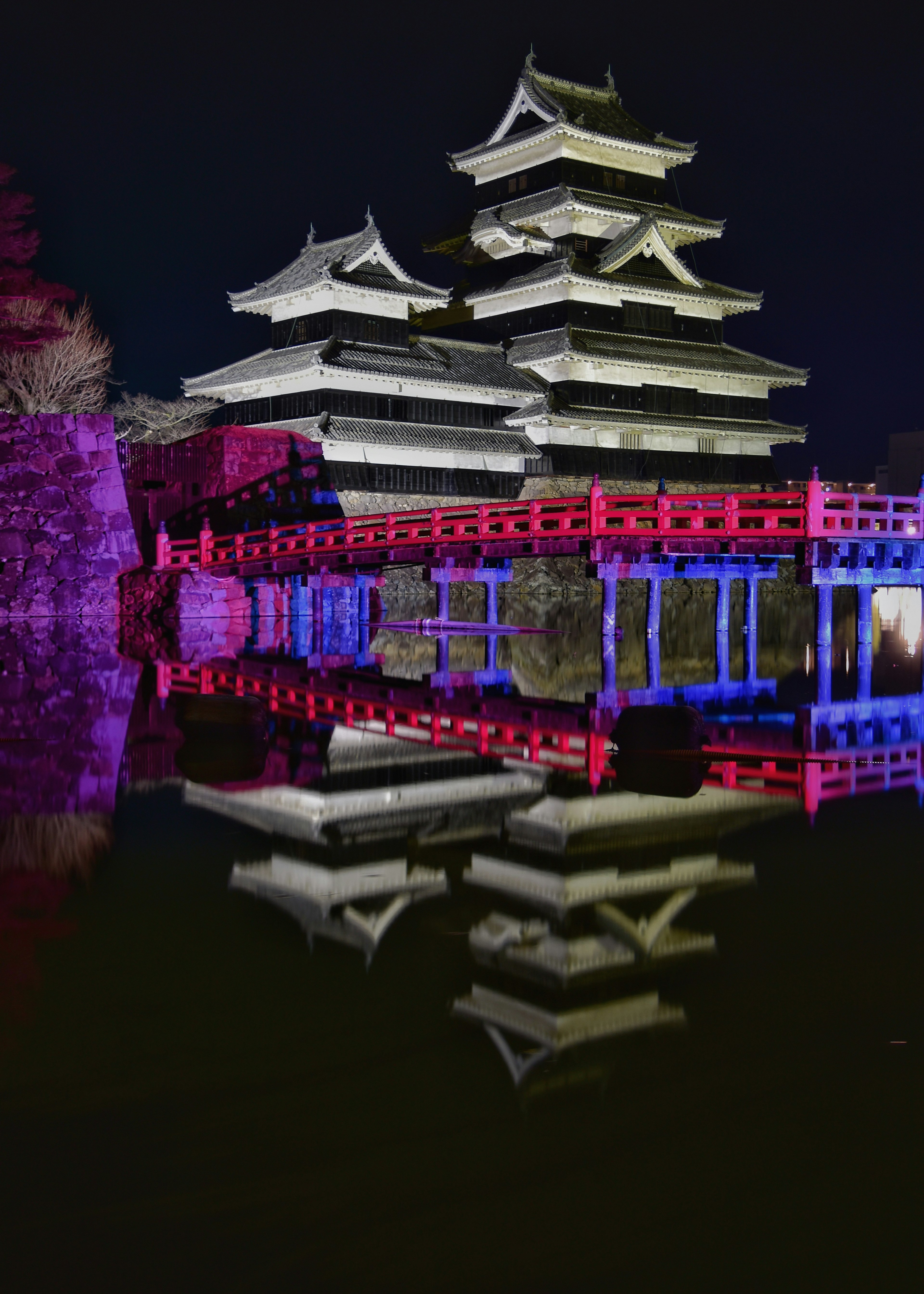 Castillo de Matsumoto de noche con reflejos coloridos y un puente rojo