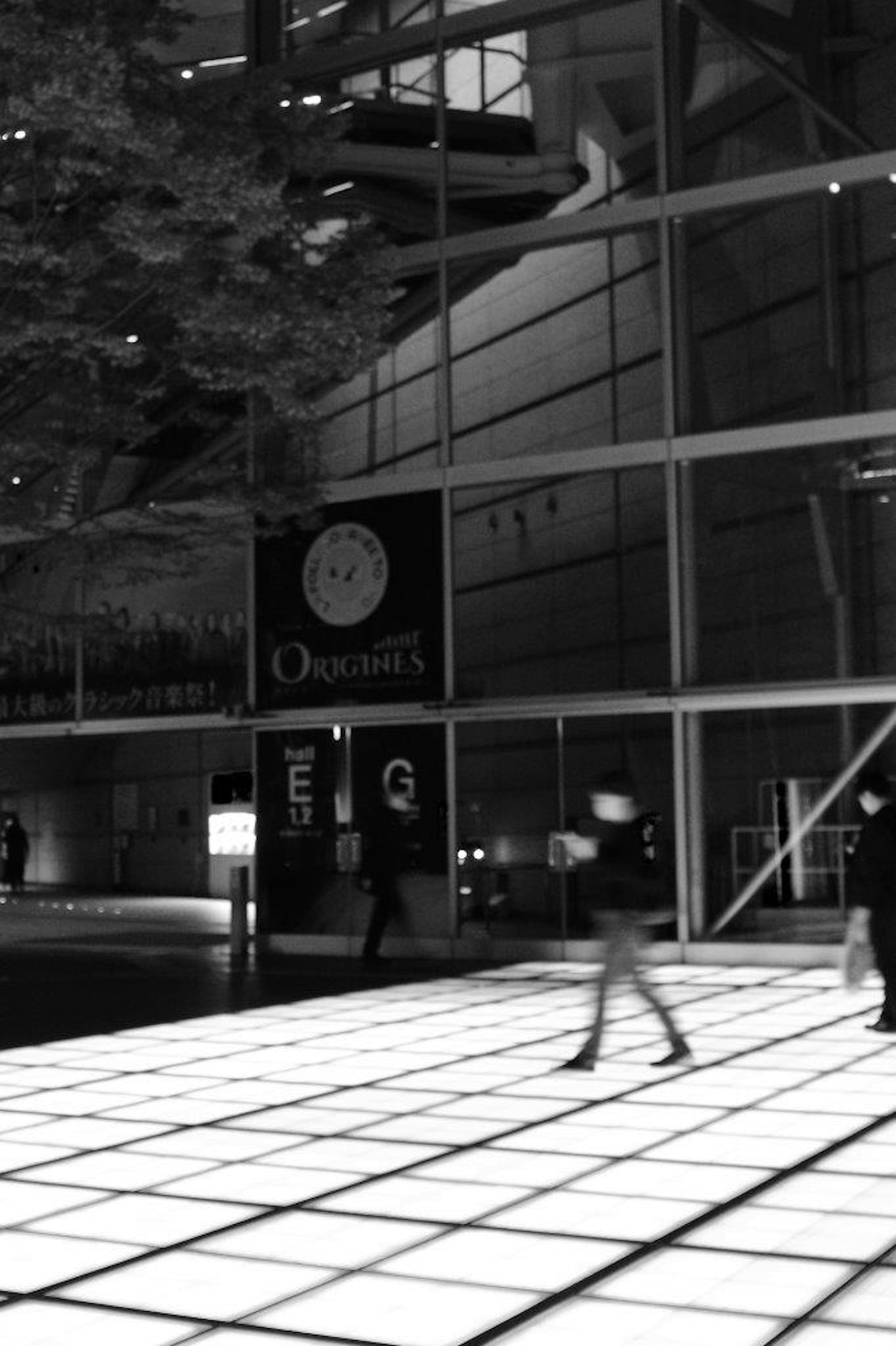 Black and white image of people walking in front of a modern building illuminated floor and glass walls