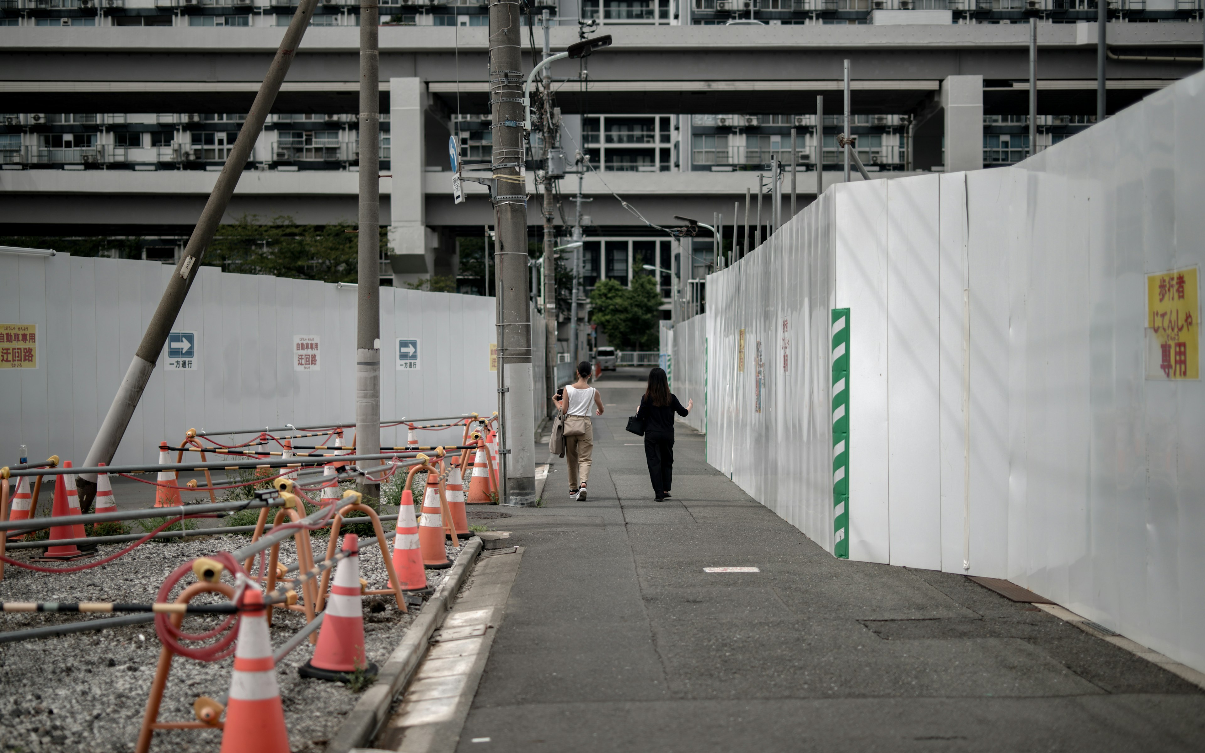 Two people walking on a sidewalk next to a construction site with white walls and orange cones