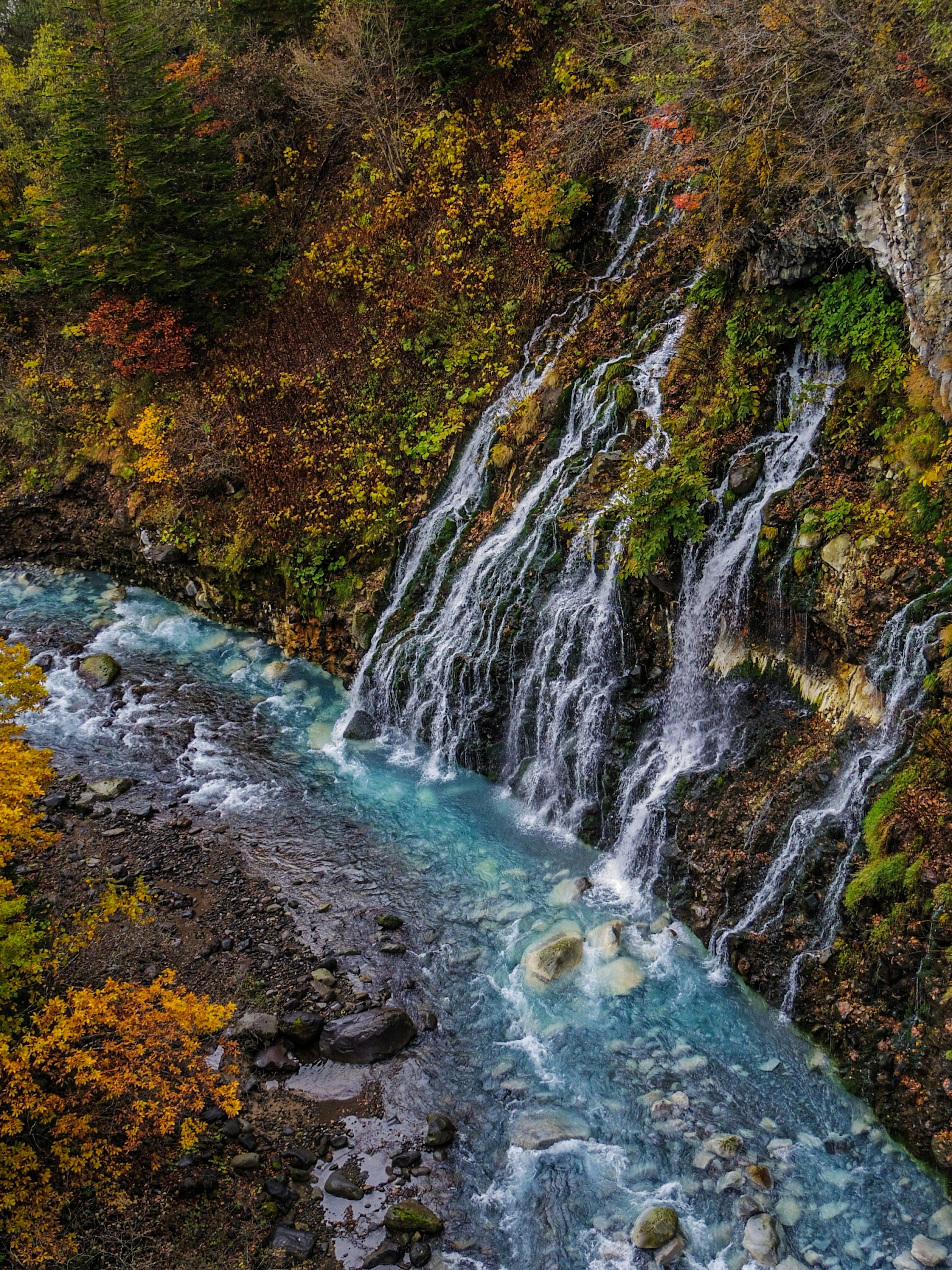 A waterfall cascading beside a clear blue river with autumn foliage