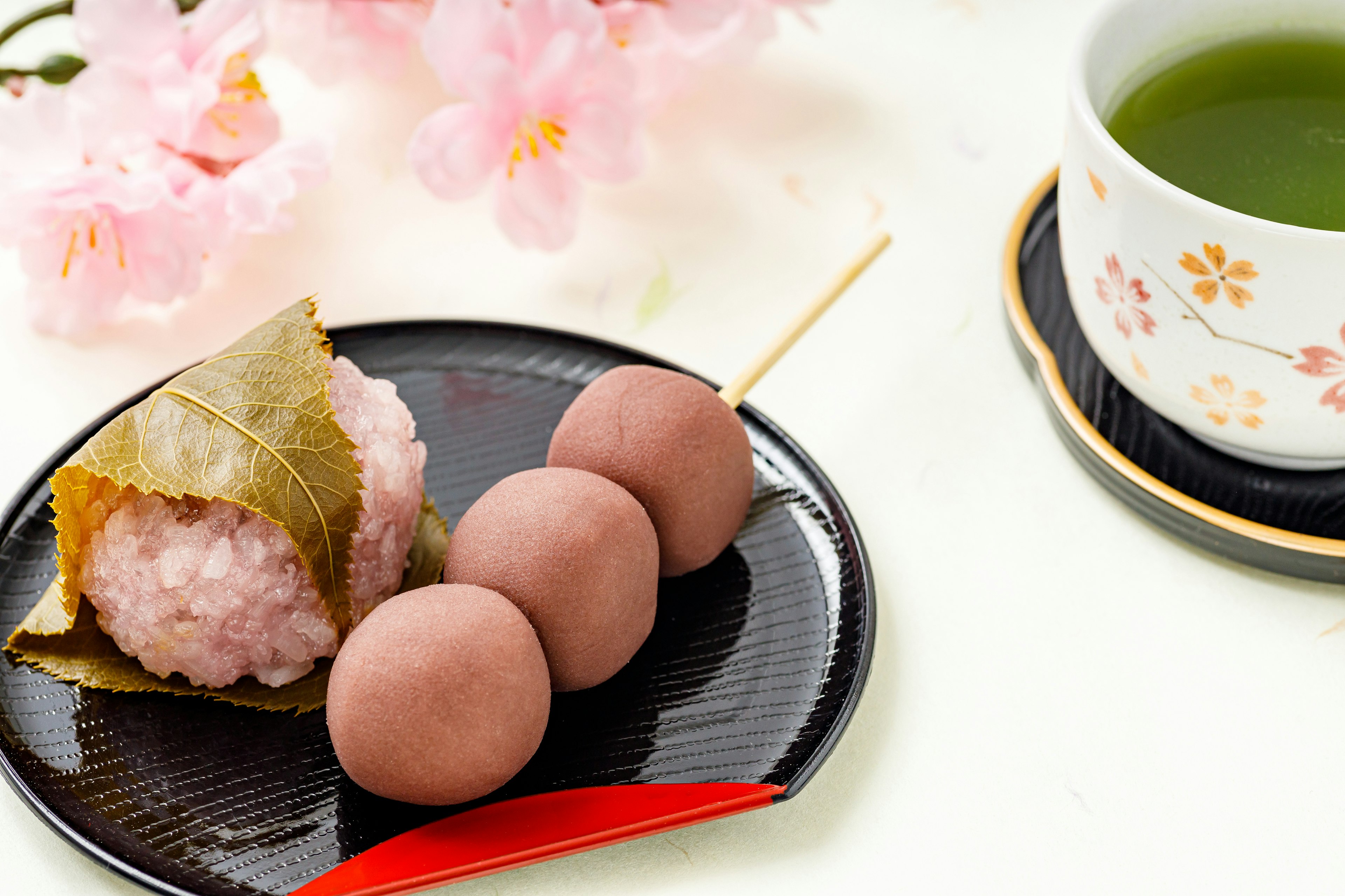 Gambar makanan manis Jepang tradisional dan teh hijau di piring hitam dengan latar belakang bunga sakura