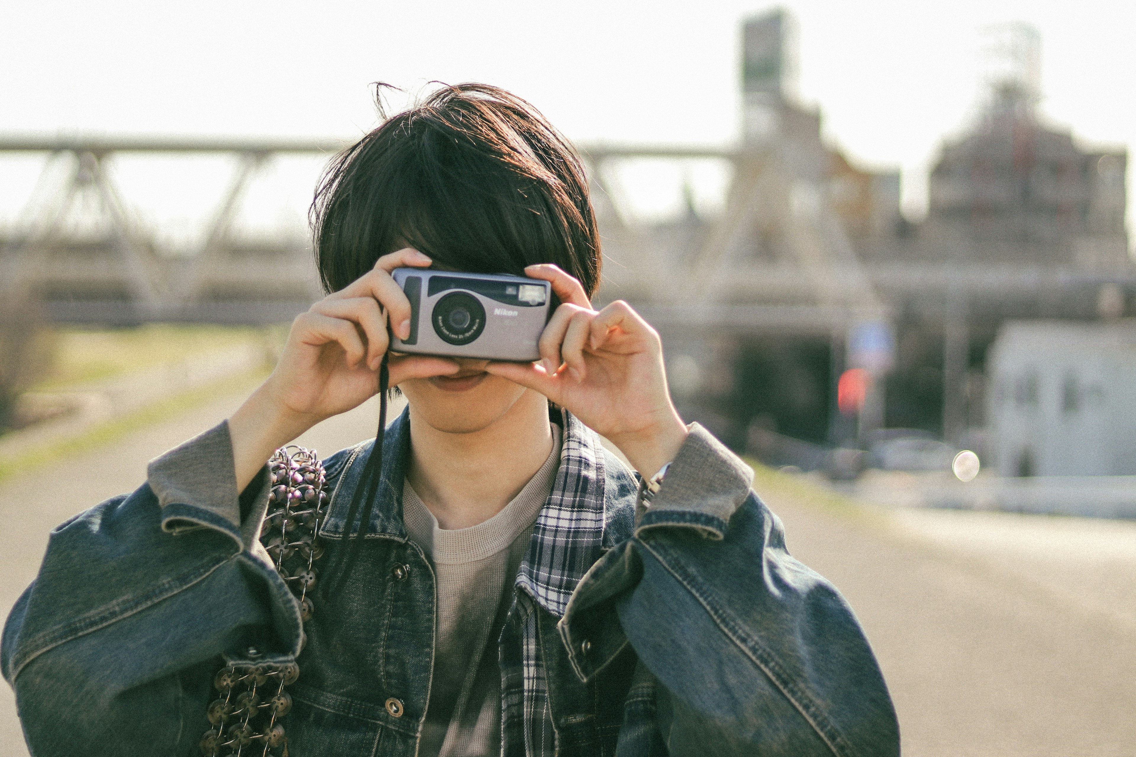 A young man holding a camera posing against a backdrop of industrial buildings