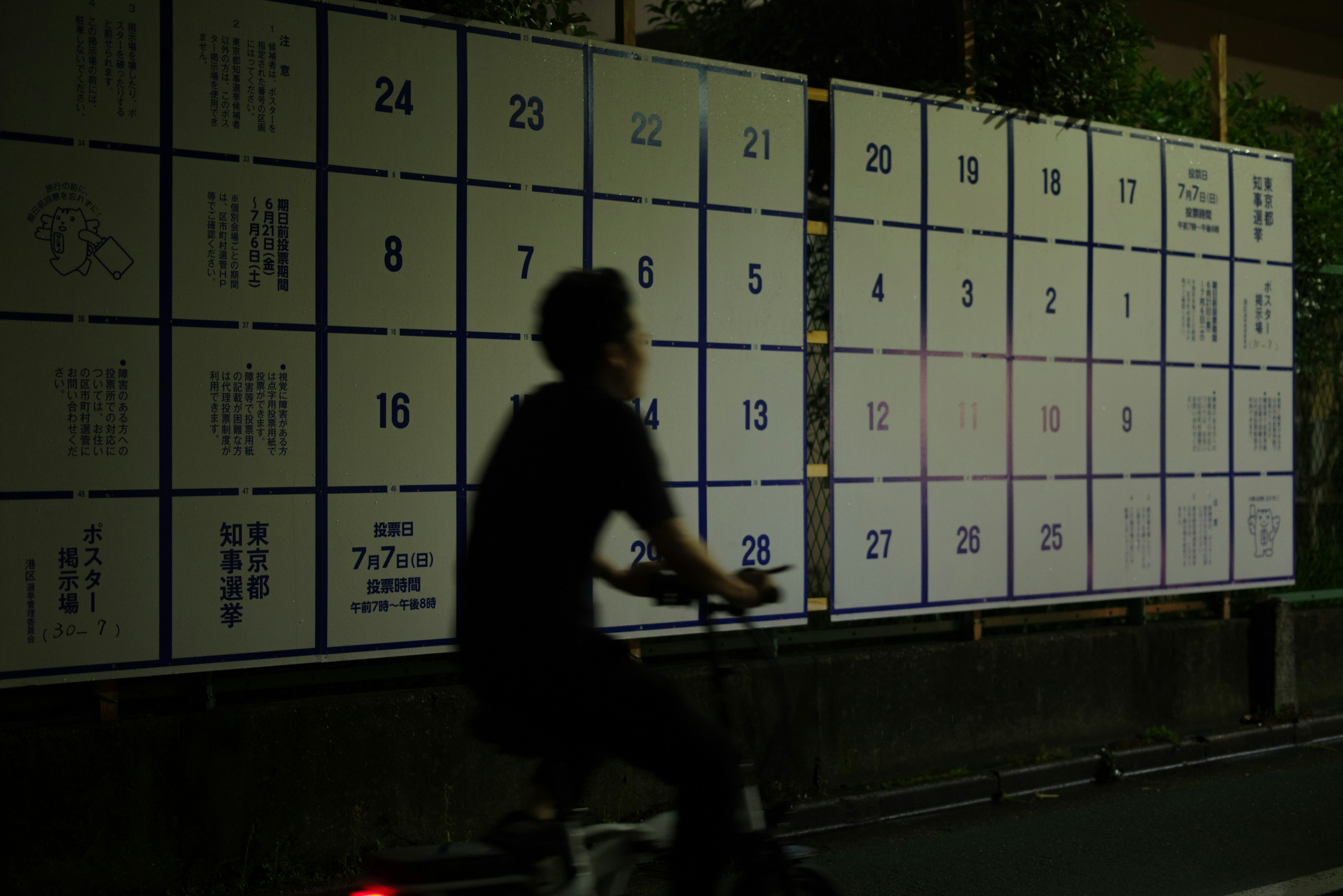 A person riding a bicycle past a wall with numbered panels at night