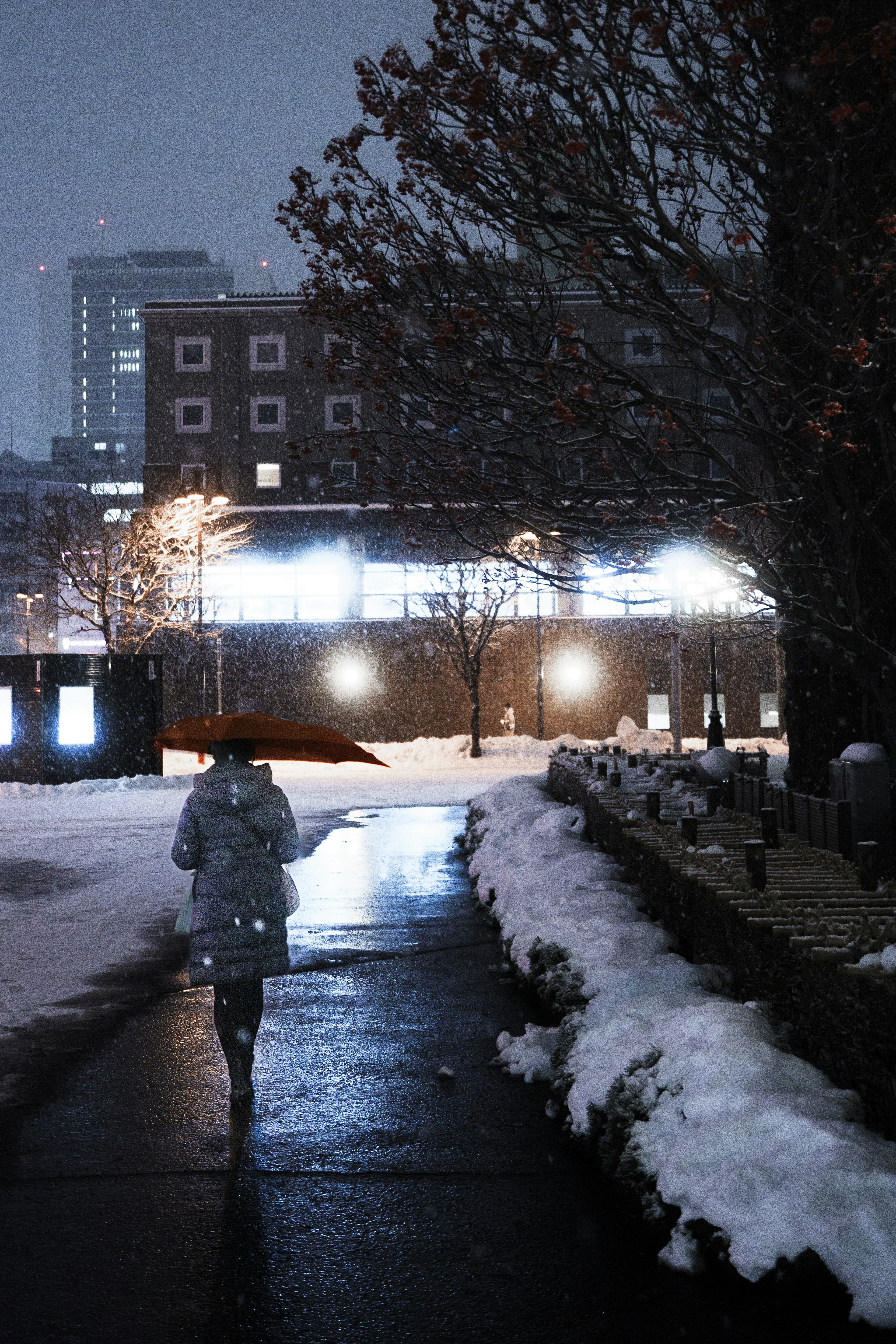 A person walking on a snowy sidewalk at night with streetlights illuminating the path