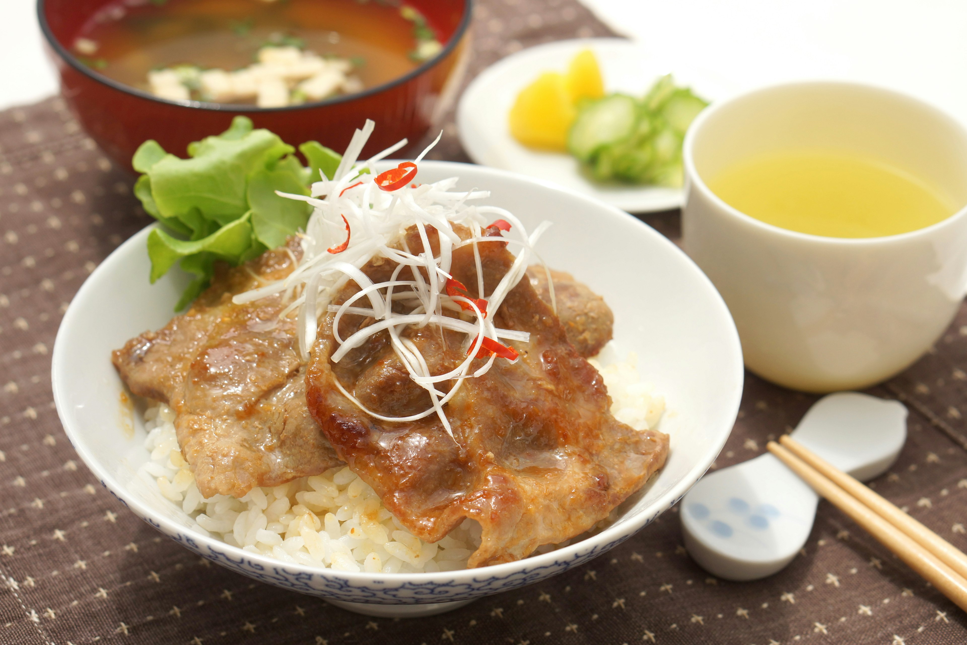 Bowl of rice topped with teriyaki pork garnished with onions and side dishes including miso soup and pickles