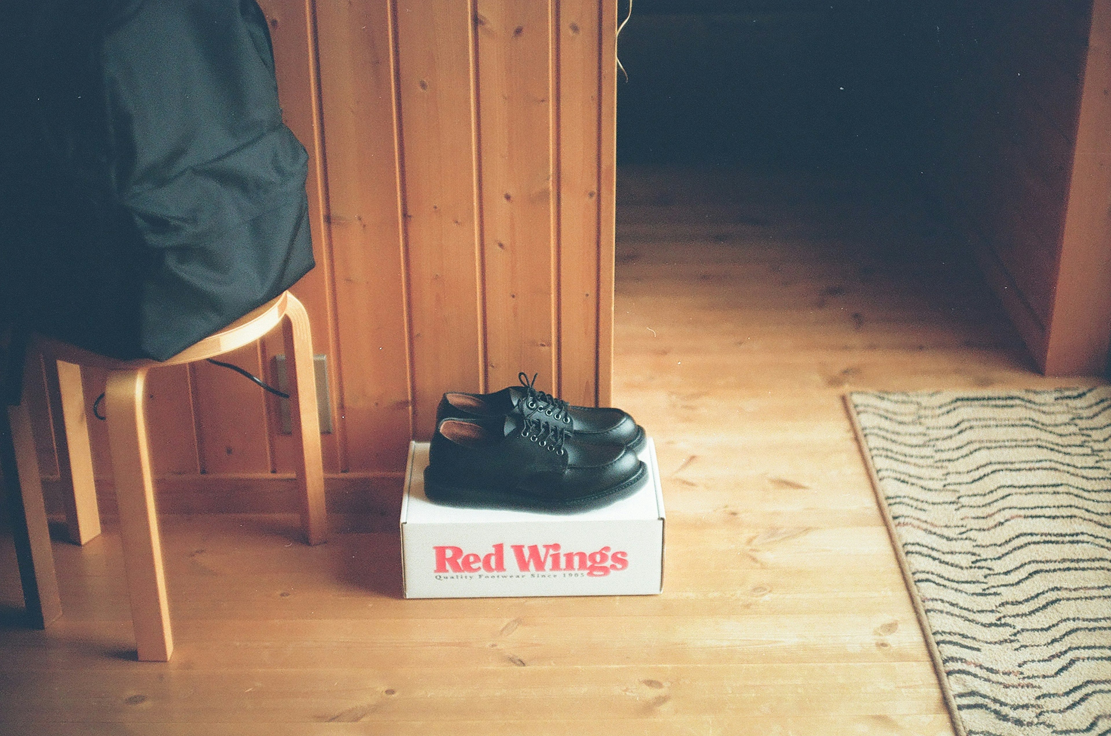 Black shoes placed on a Red Wings box in a room with wooden flooring