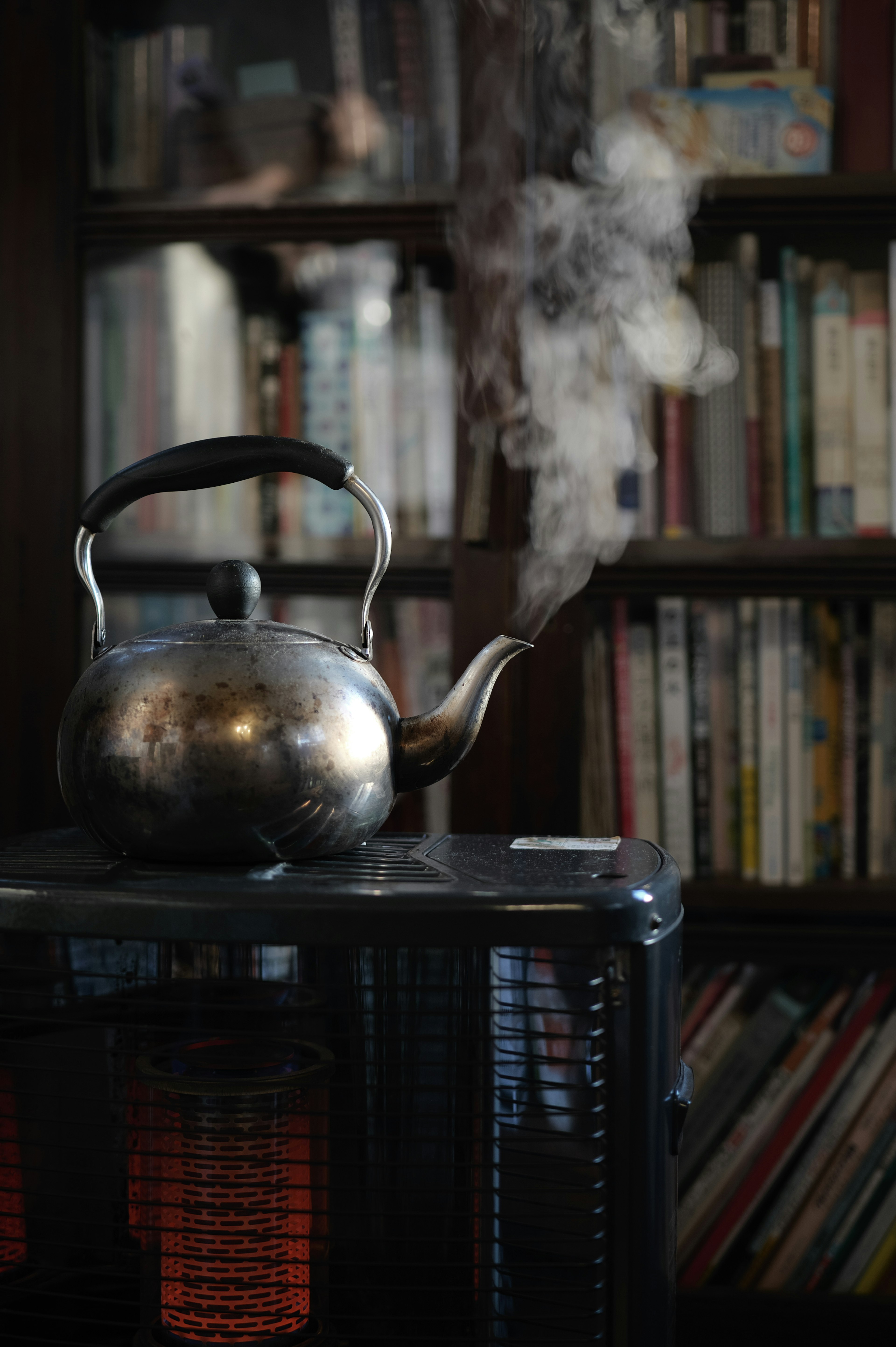 Silver kettle emitting steam on top of a heater with a bookshelf in the background