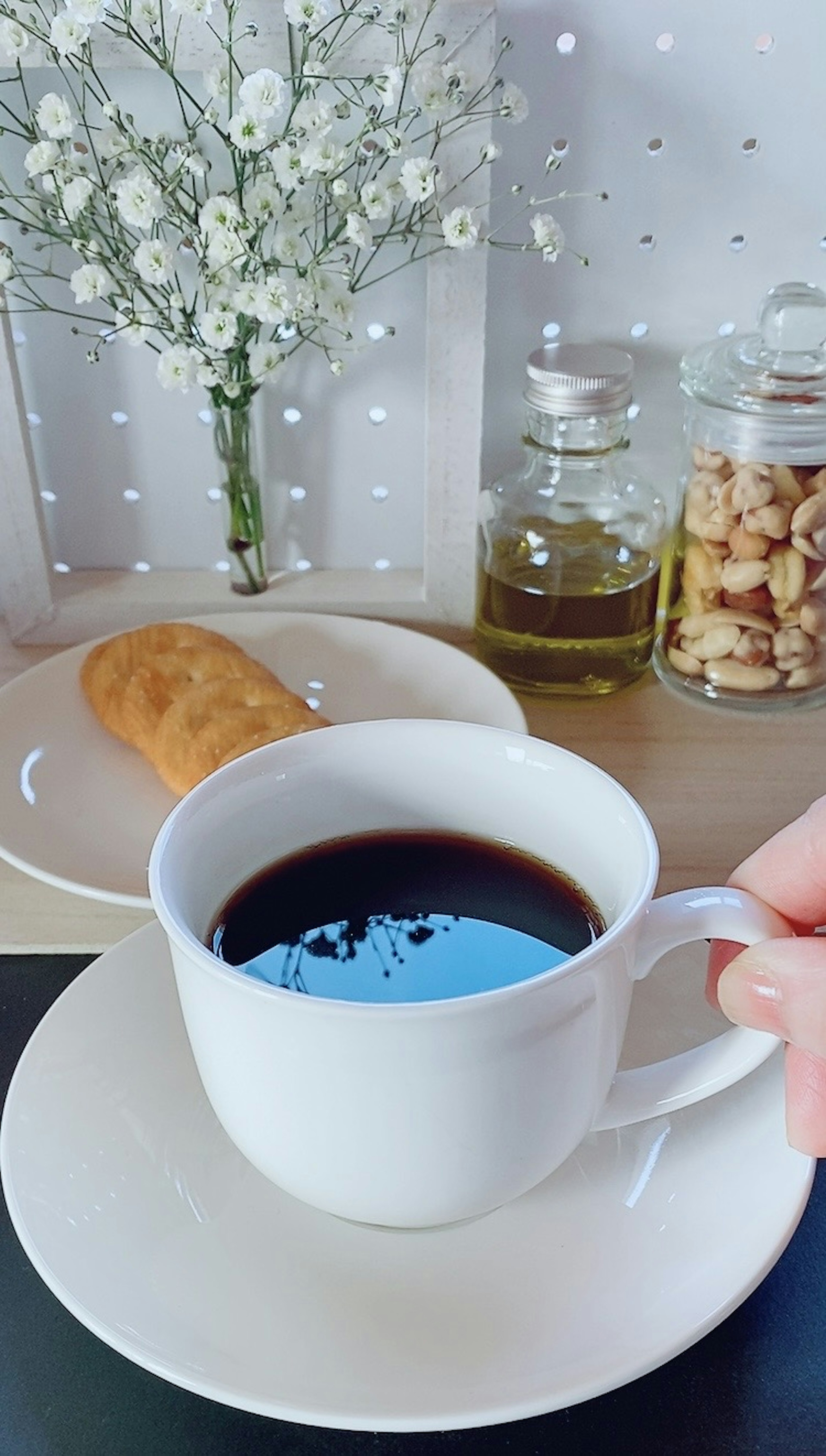 Una mano sosteniendo una taza blanca de café negro junto a un plato de pasteles y flores decorativas