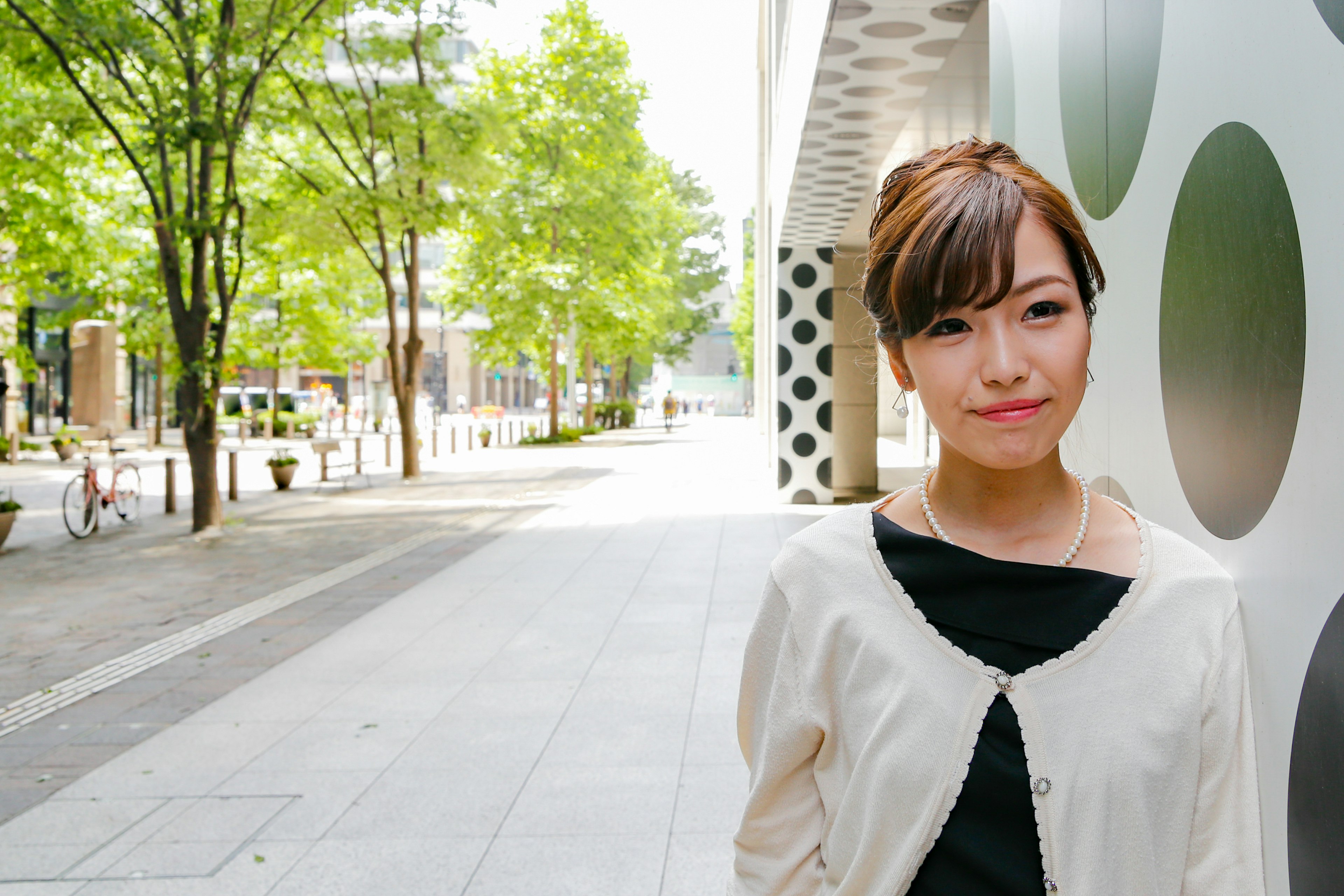 A woman smiling in an urban setting with green trees and modern buildings in the background