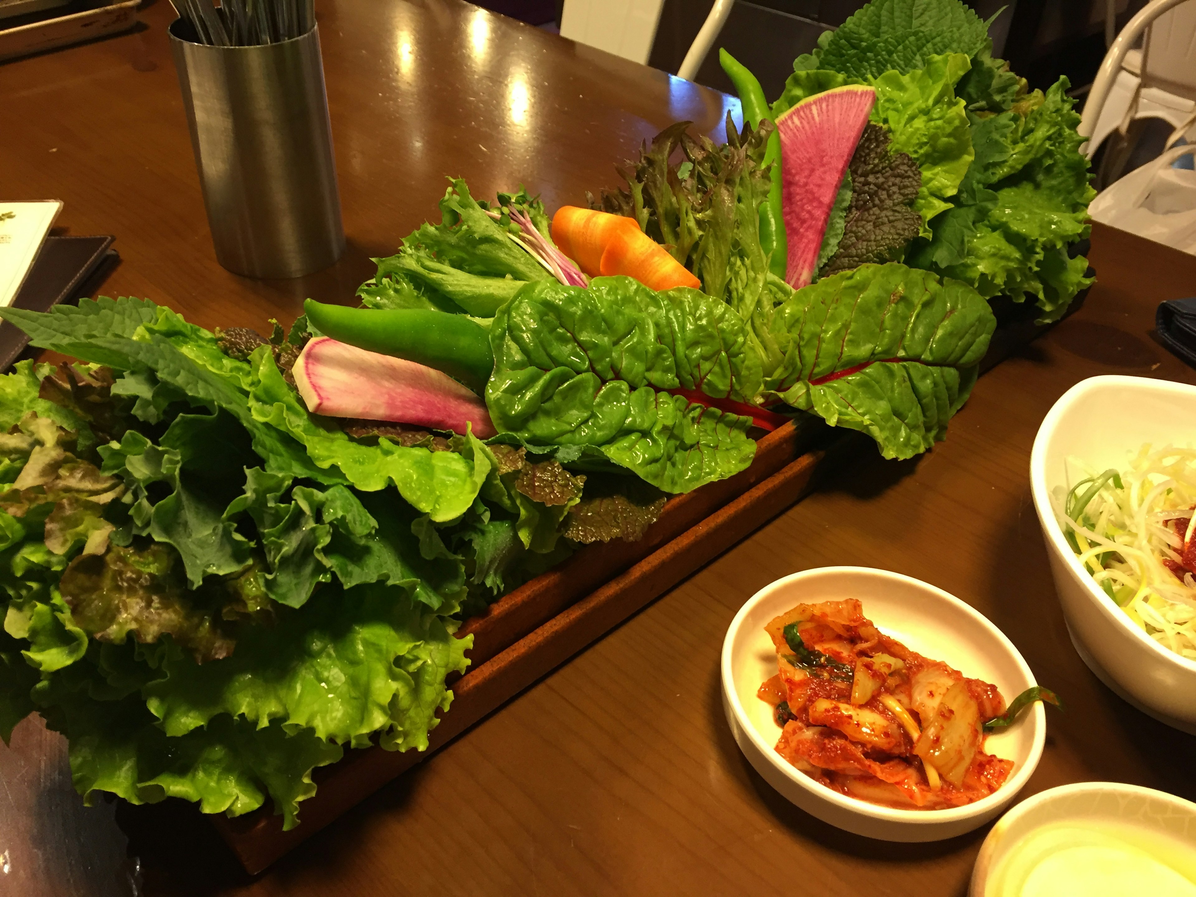 Una bandeja de verduras frescas que incluye lechuga, rábanos y zanahorias con un lado de kimchi