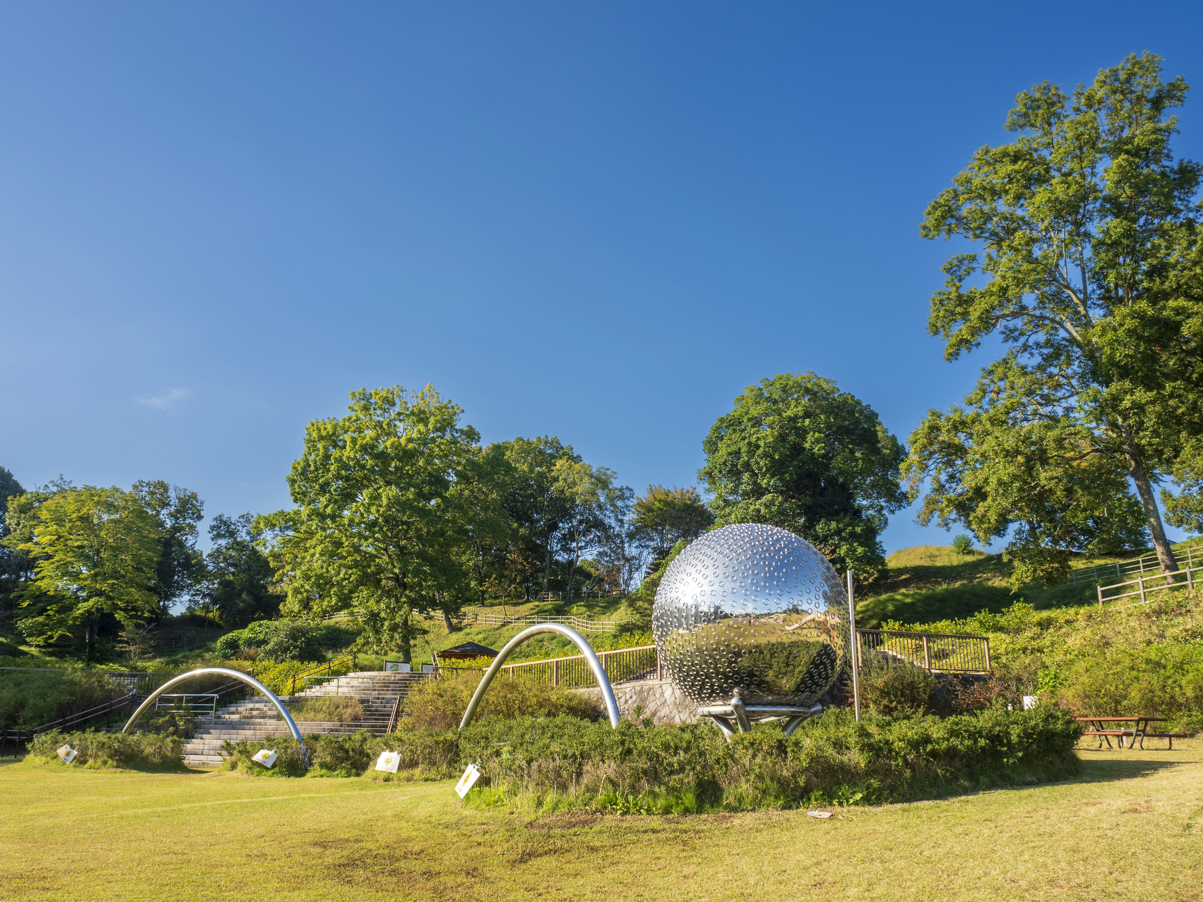 青空の下にある鏡のような球体とアーチ状の彫刻が配置された公園の景観