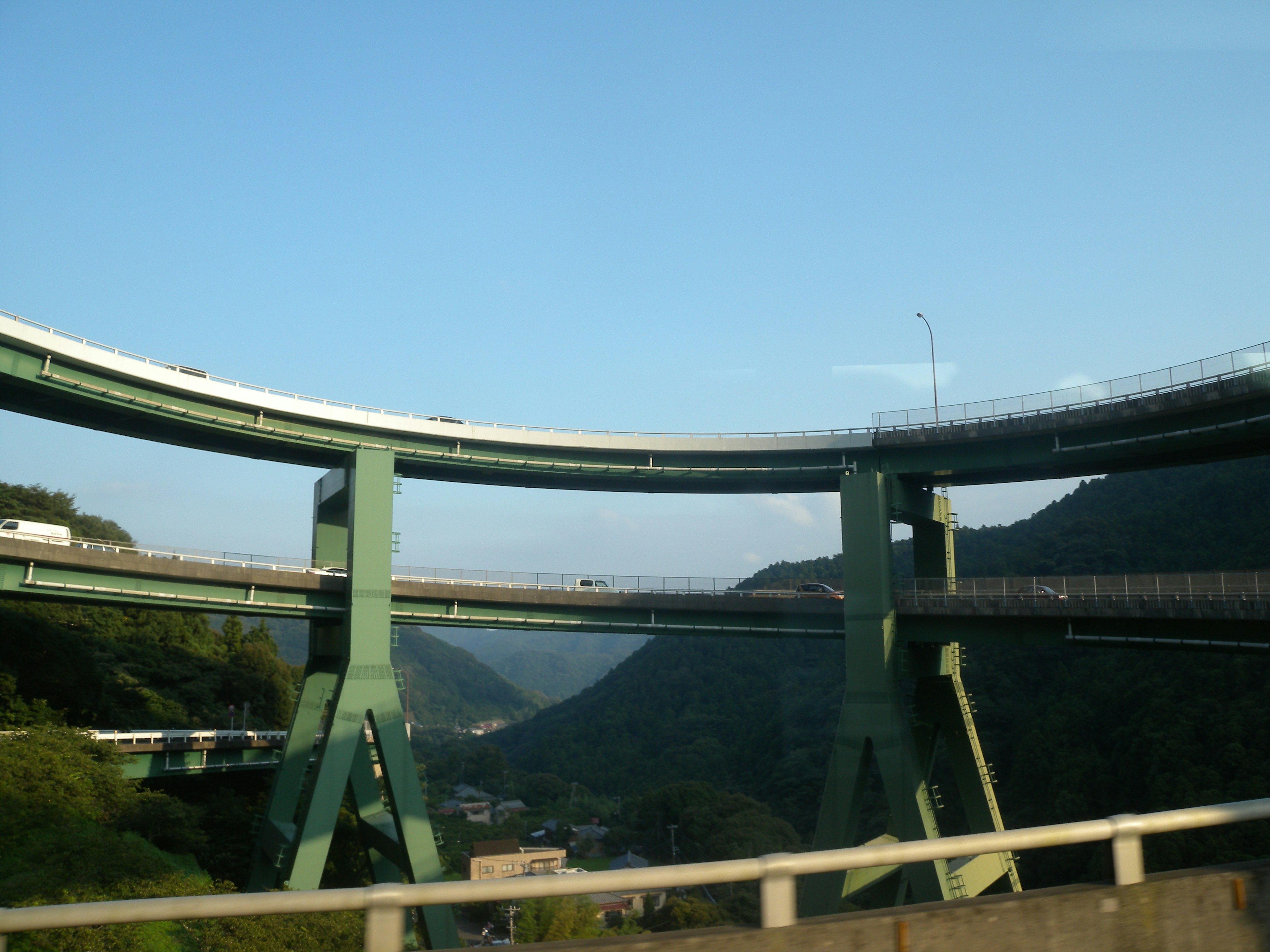 Jalan tol hijau melengkung melalui pemandangan pegunungan dengan langit biru