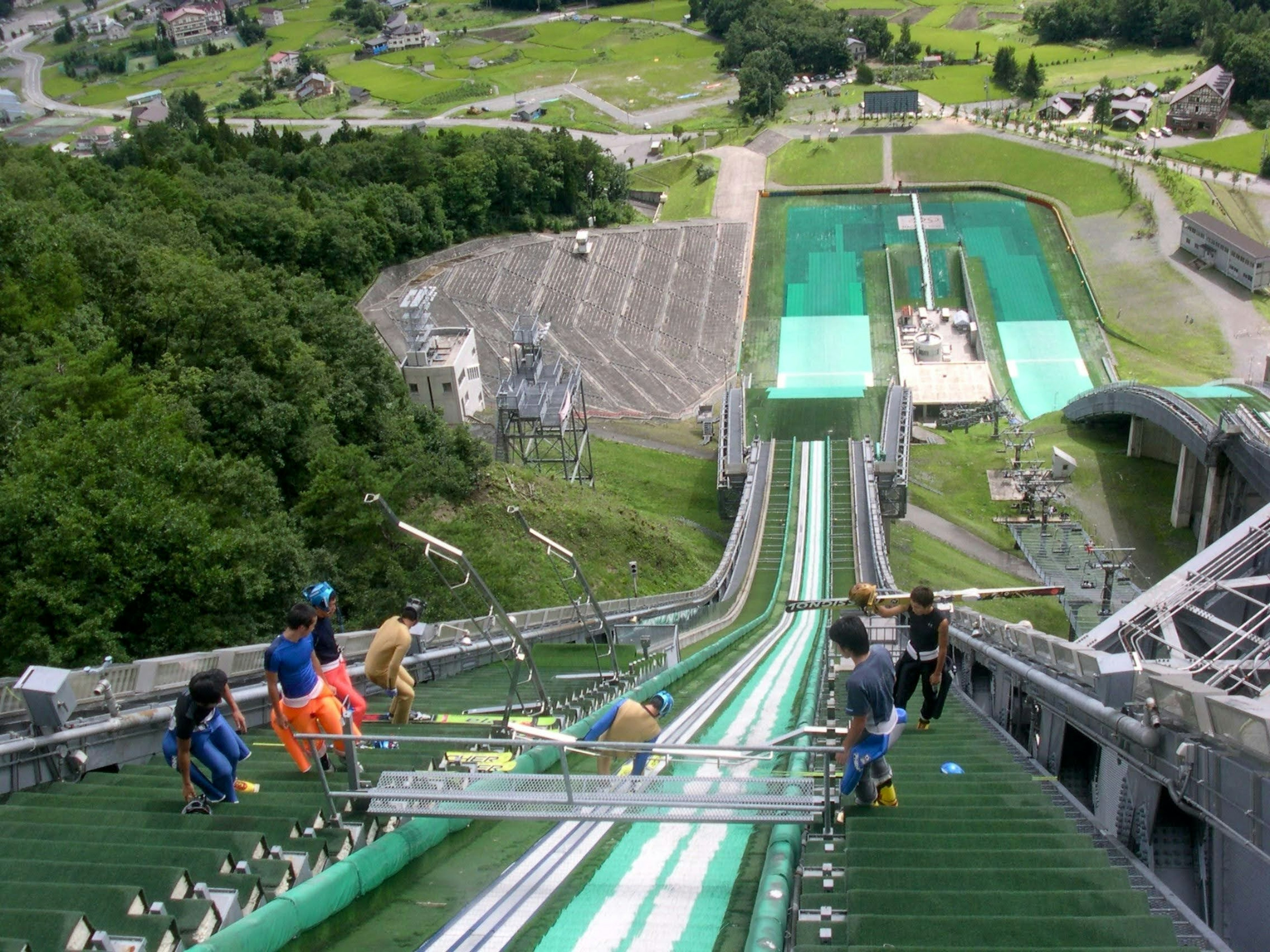 Blick von der Spitze einer Skisprungschanze mit Athleten, die sich auf der grünen Rampe vorbereiten