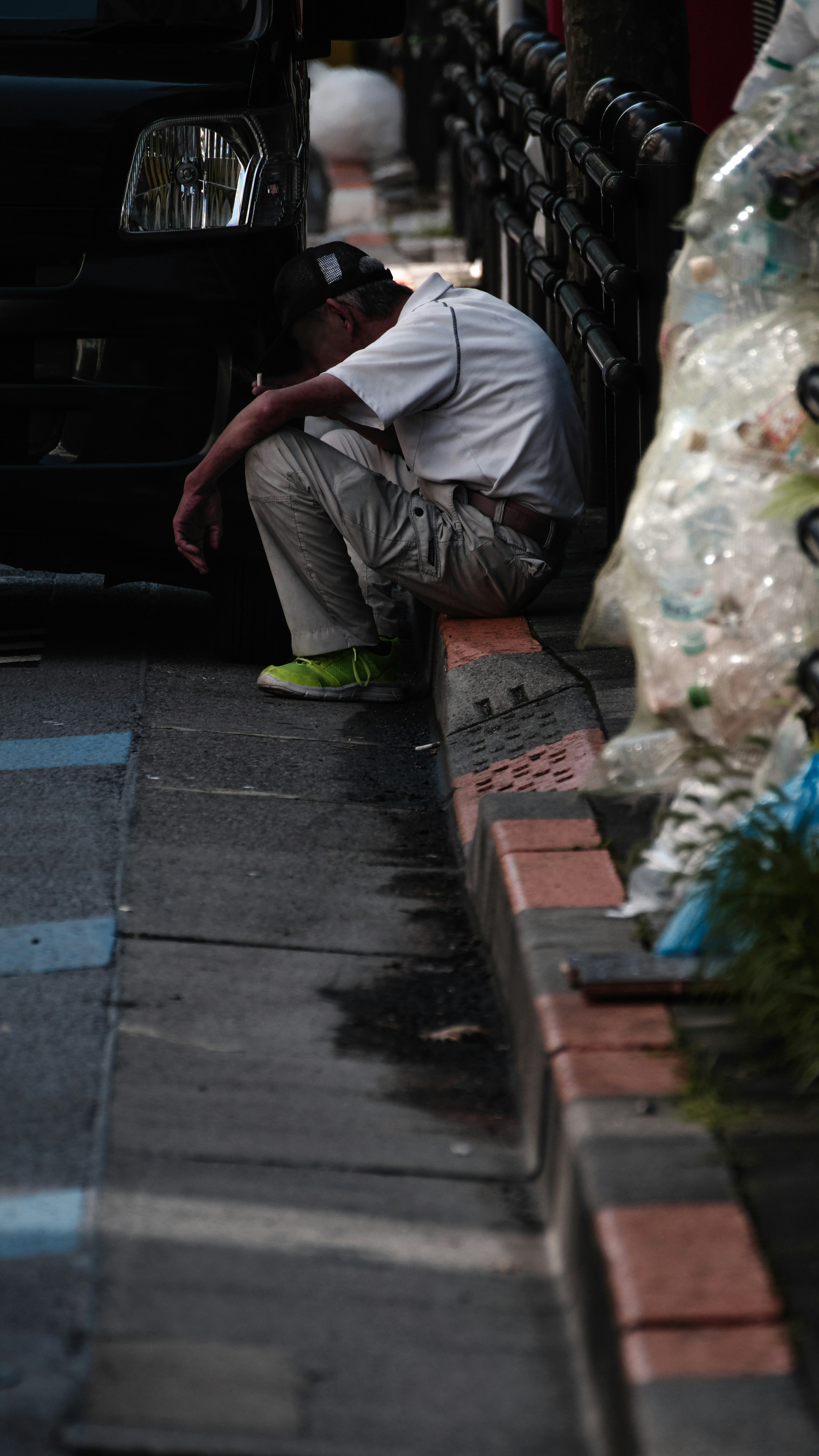 Un hombre sentado al borde de la carretera con pantalones cortos y una camiseta blanca zapatos de colores cerca de la basura con una expresión contemplativa