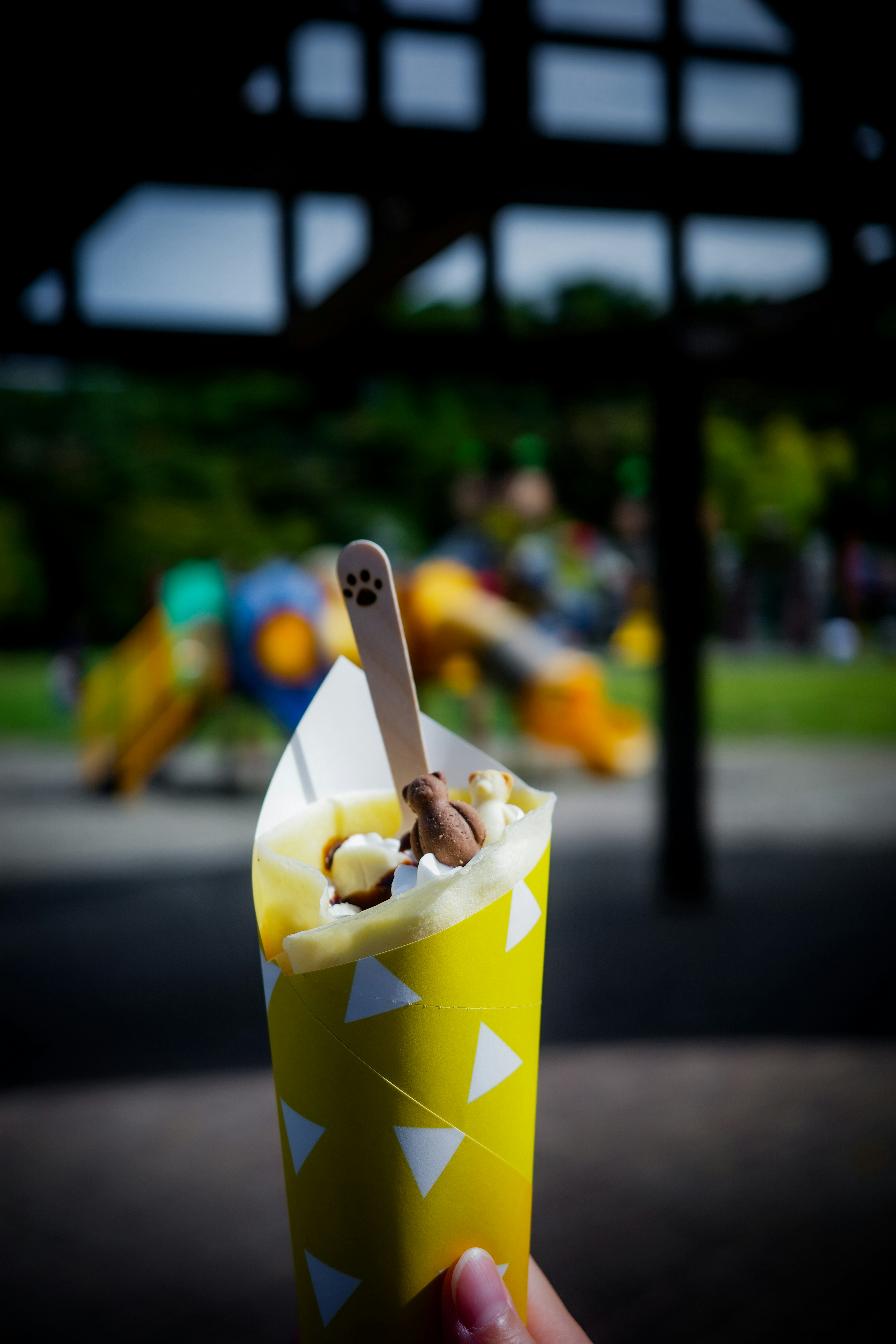 Primo piano di gelato in un cono di carta giallo a motivi triangolari con un cucchiaio di legno sfondo sfocato di un parco giochi