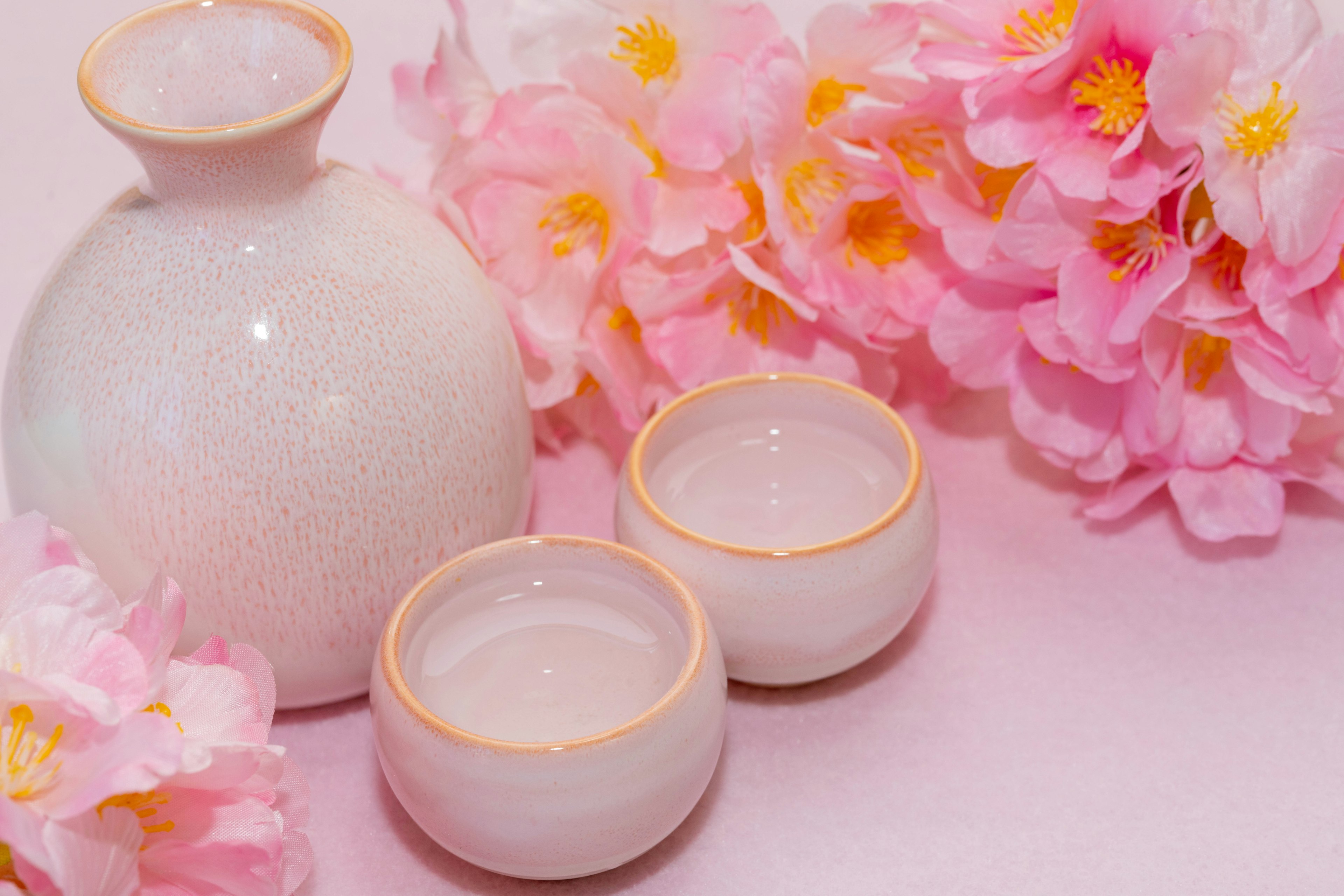 White ceramic sake bottle and two cups on a soft pink background surrounded by cherry blossoms