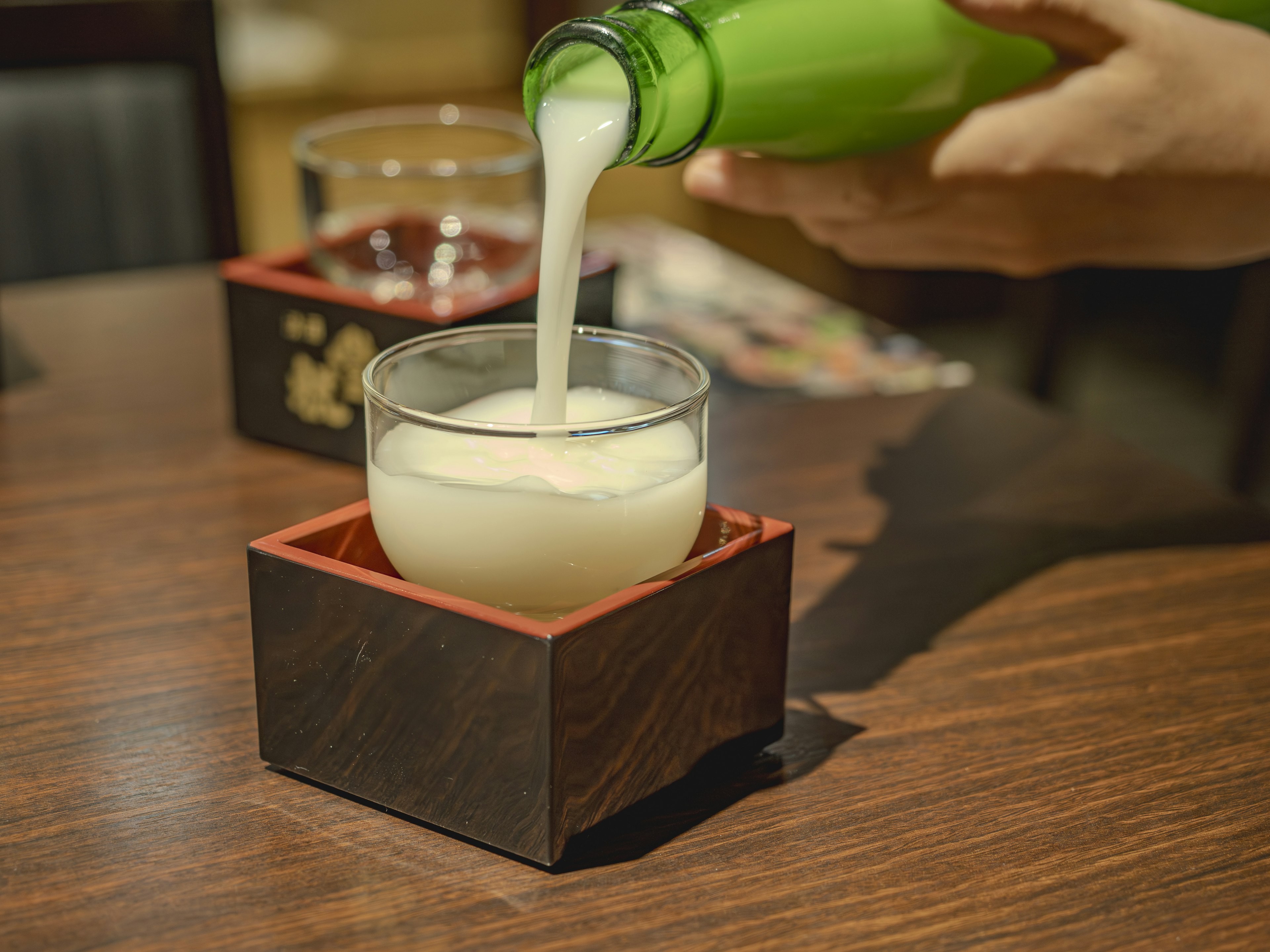 Einschenken eines weißen Getränks aus einer grünen Flasche in ein Glas auf einem Holztisch