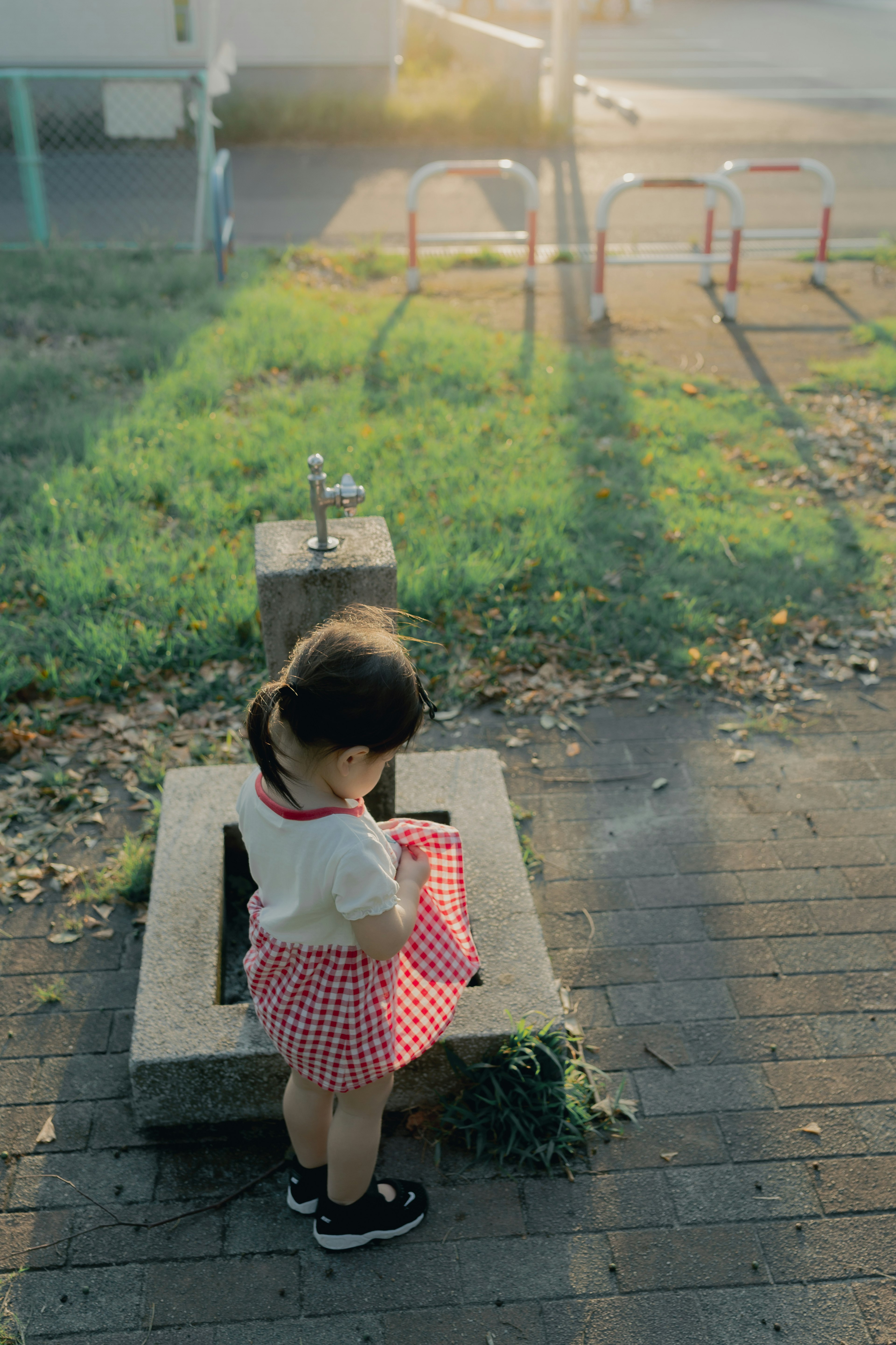 Una niña pequeña está de pie frente a una fuente en un parque, vistiendo una falda a cuadros rojos